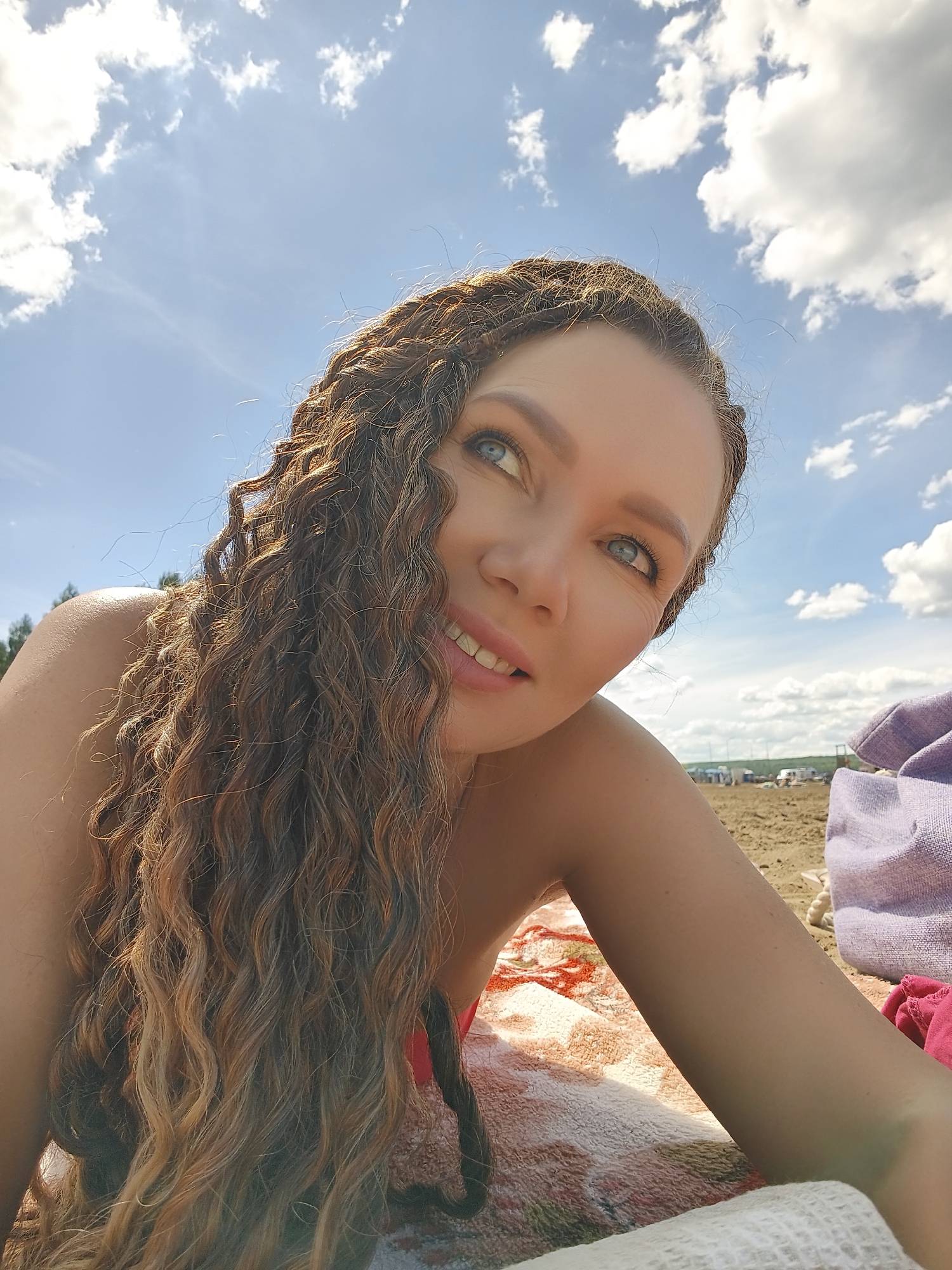 A young woman with long, curly brown hair is lying on a beach. She is looking up and to her right, with a slight smile on her face. Her eyes are blue and her skin is fair. The sun is shining brightly, and there are some clouds in the sky. The background shows a sandy beach with some vegetation in the distance. She is wearing a red bathing suit and lying on a colorful blanket. There are also some bags and towels on the sand next to her. The overall mood is relaxed and happy, suggesting a day at the beach.