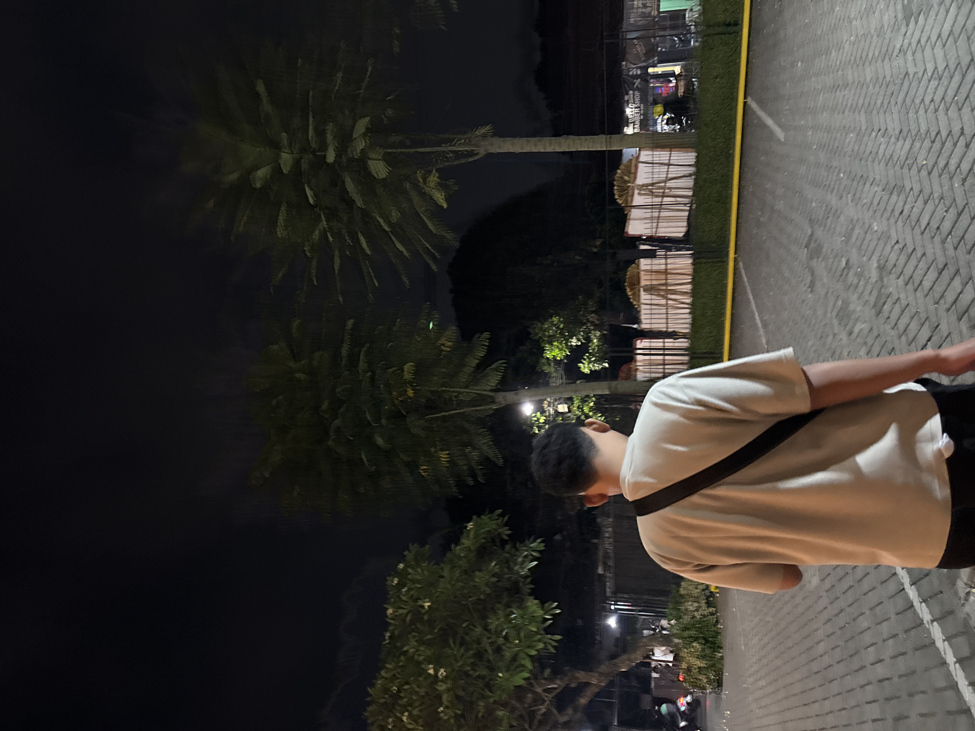 The image shows the back of a person wearing a light-colored t-shirt and dark pants, with a black strap across their chest. They are standing in what appears to be an outdoor parking lot paved with grey interlocking bricks, marked with white and yellow lines. In the background, there are several trees, including palm trees, and dimly lit structures, some of which appear to be small shops or stalls. The scene is set at night, with the sky being dark. One of the structures in the background has signage that reads "BELLO BARBER SHOP." The overall environment suggests an urban or semi-urban setting in South Tangerang, Indonesia, possibly a commercial or recreational area after dark.