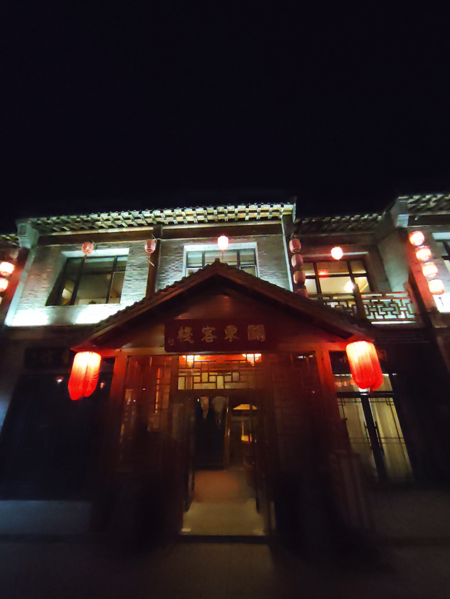 This image depicts a nighttime scene in the city of Lindian, China, featuring a traditional Chinese-style building adorned with red lanterns. The building appears to be a commercial establishment, possibly a restaurant or a guesthouse, given the ornate architectural details and signage. The facade is constructed with brick and features multiple levels with windows and decorative elements. Red lanterns, a common symbol of good fortune and celebration in Chinese culture, are hung at various points, casting a warm, inviting glow. The entrance to the building is framed by a more elaborate wooden structure, further enhanced by the illumination of more lanterns. The overall impression is one of cultural richness and festive ambiance, typical of traditional Chinese urban landscapes during evening hours.