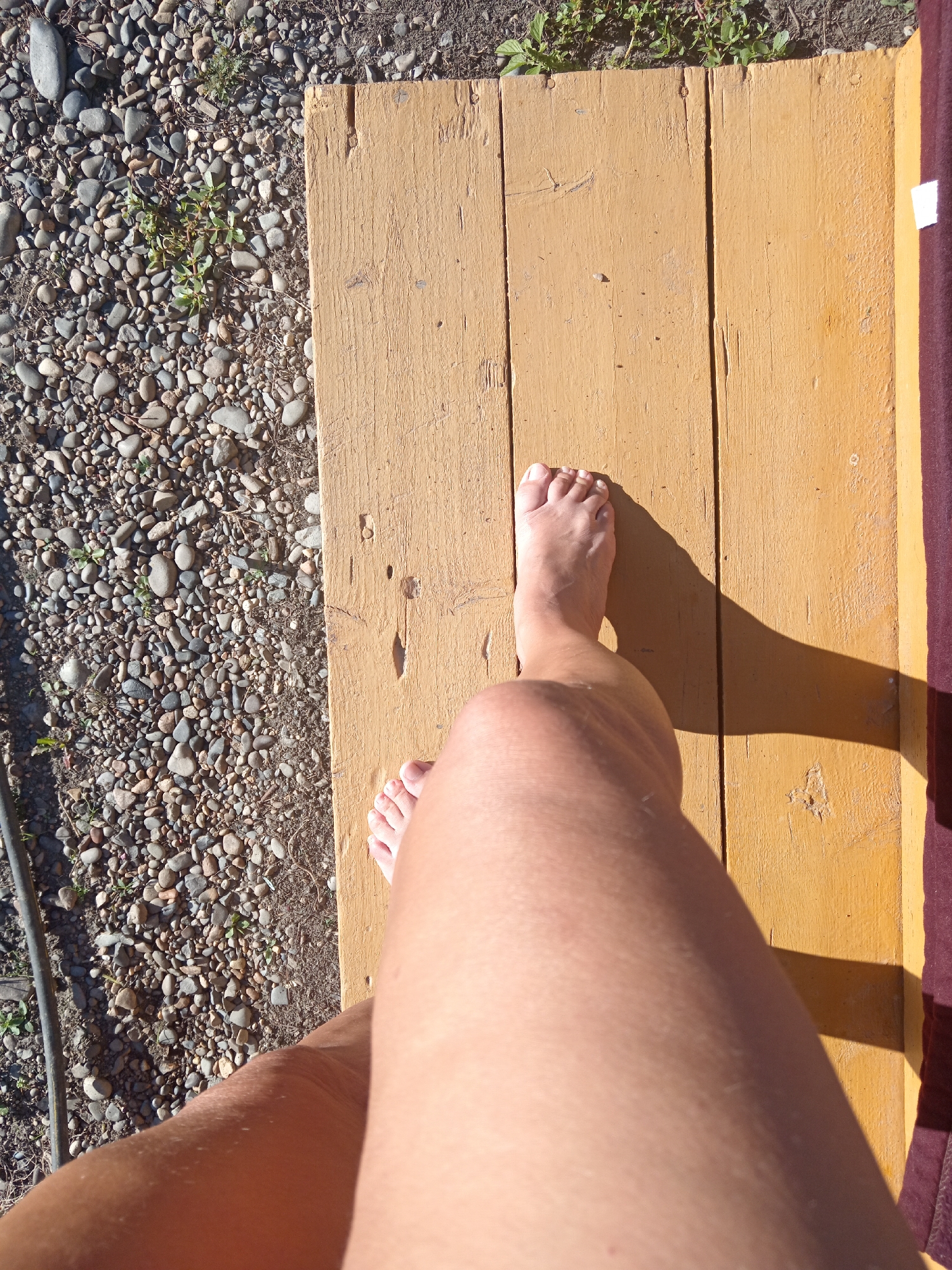 The image shows a person's left foot and lower leg standing on a yellow wooden surface. The wooden surface appears to be part of a deck or porch, composed of several planks. To the left of the wooden surface is an area covered in small, multicolored rocks and some sparse green vegetation. A dark, coiled cable is visible on the far left edge of the image, partially obscured by the rocks. The light suggests it is daytime, casting a shadow of the foot and leg onto the wooden surface. The context of Novopokrovka, Russia, suggests an outdoor setting, possibly a residential area given the decking and stony ground.