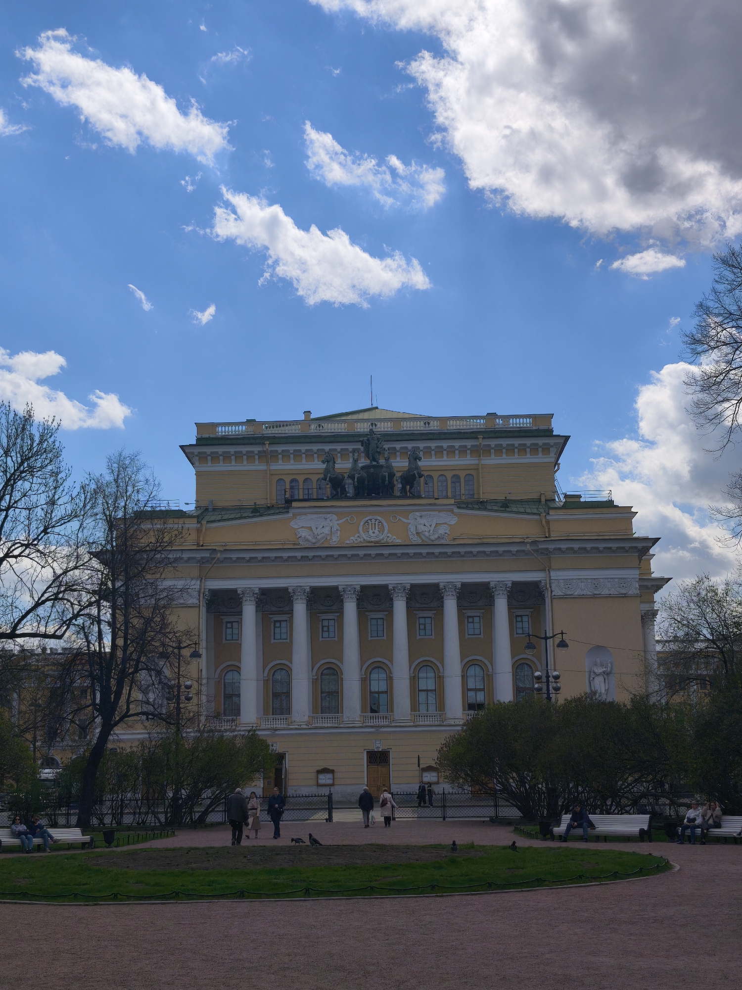 The image shows the Alexandrinsky Theatre in Saint Petersburg, Russia. The building is a light yellow color with white columns and a statue on top. There is a park in front of the theatre with trees, benches, and people. The sky is blue with white clouds. Some people are sitting on the benches, while others are walking through the park. Pigeons are also visible on the ground.