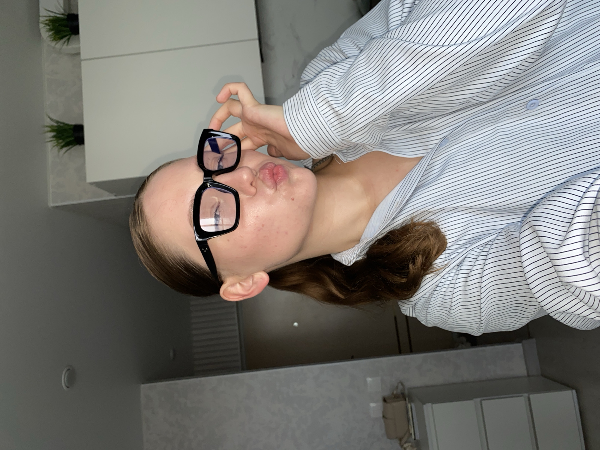 A young woman with light skin and brown hair tied in a ponytail is posing for a selfie in a room. She is wearing black-rimmed glasses and a light blue and white pinstriped button-up shirt. Her lips are pursed in a kiss pose. The background shows a white cabinet with two artificial plants on top, a gray textured wall, and a white dresser. The lighting suggests an indoor setting, possibly a home. The image is a close-up, focusing on the woman's face and upper body. There is no indication of the location being Izhevsk, Russia.