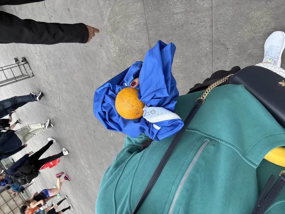From a high-angle perspective, the scene captures a person standing on a gray concrete surface. They are wearing a green jacket and white sneakers and a black bag. Resting on the jacket is a blue shirt with a round, orange fruit nestled inside. In the background, other people are walking in various directions. The overall composition creates a snapshot of a moment in a busy public space.