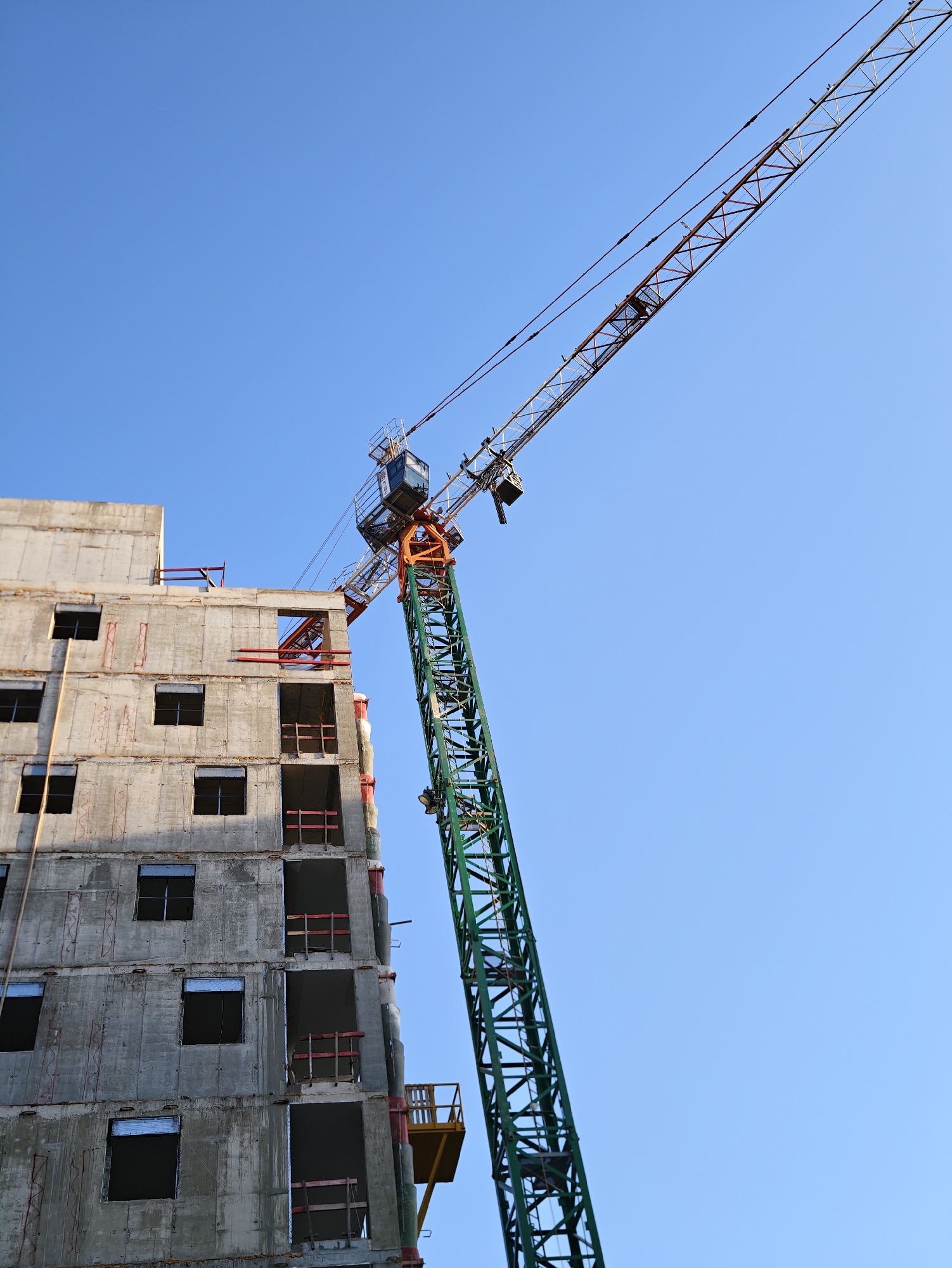 A construction crane is actively engaged in building a multi-story concrete structure under a clear blue sky. The building, in its early stages of construction, has open windows and visible scaffolding. The crane's green tower rises vertically, with its long jib extending diagonally across the frame towards the right. A small operator's cabin is visible at the top of the crane's tower. A yellow platform, likely for material transport, is attached to the crane's tower near its base. The scene is typical of ongoing urban development and construction projects. The location of this construction site is the city of Petah Tikva, Israel.