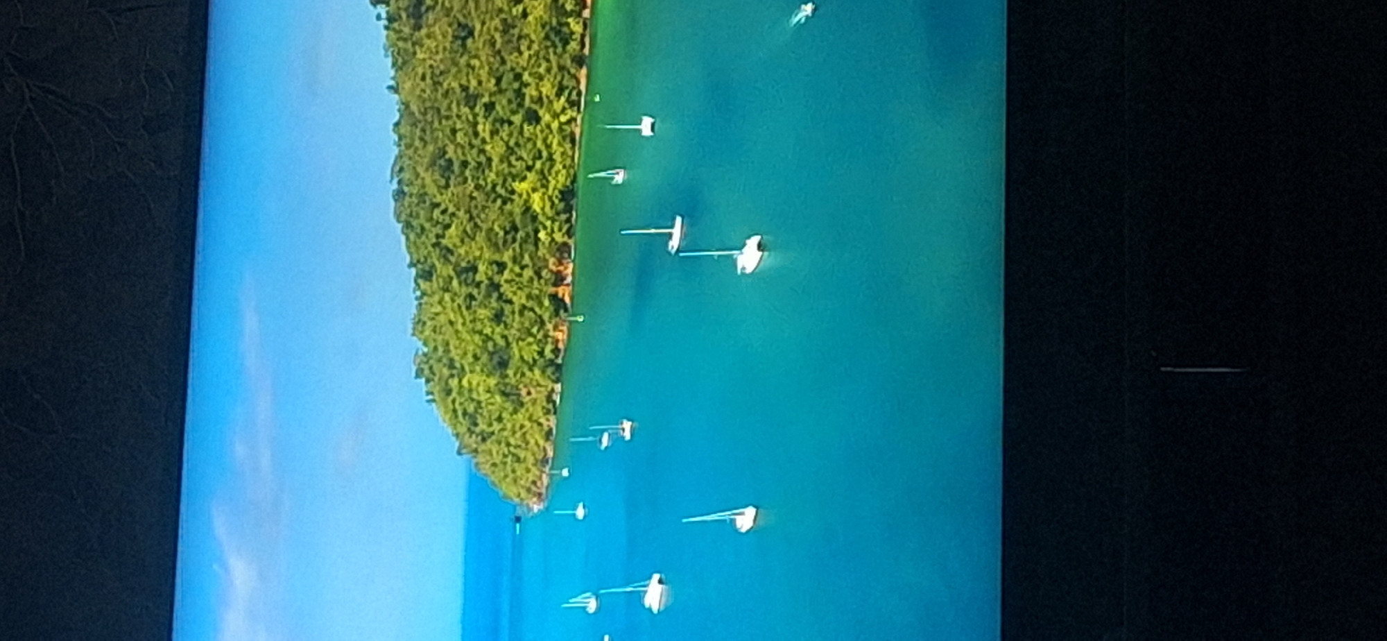 This is an aerial view of a bay with several sailboats docked. The water is a vibrant turquoise color, and a lush green island is visible in the background. The sky is clear and blue. There are no people visible in the image. The scene does not depict any locations in the city of Bila Tserkva, Ukraine, as it shows a tropical or subtropical coastal environment.