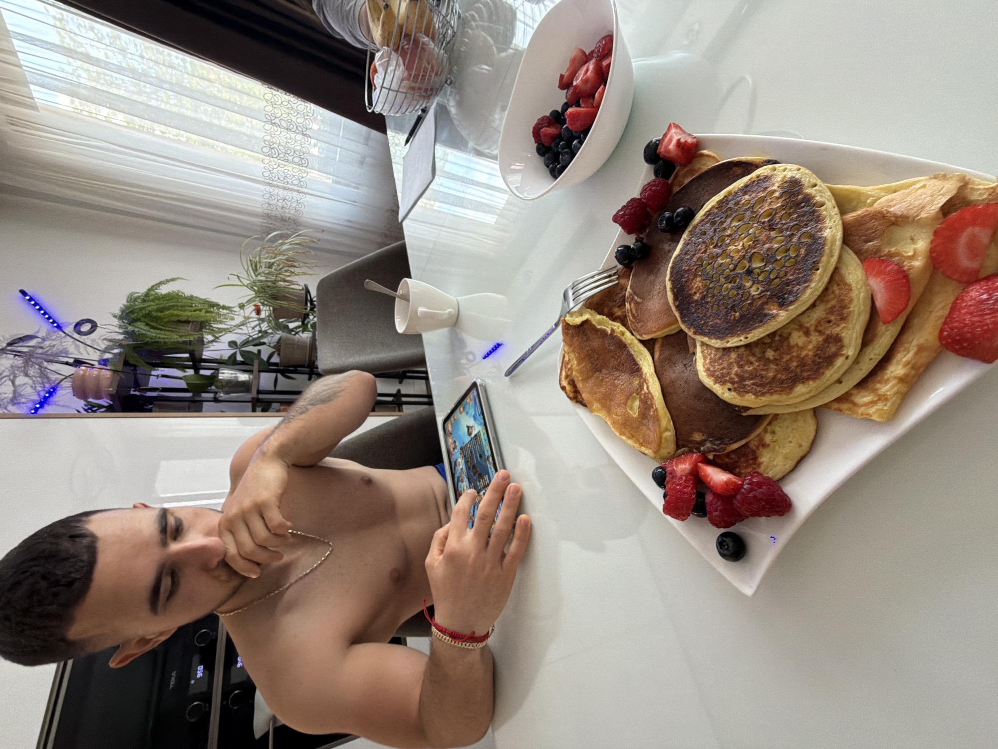 A shirtless man with a gold chain and bracelets is seated at a white table, looking at a tablet. He is located in an indoor environment. On the table is a plate of pancakes and crepes garnished with strawberries, raspberries, and blueberries, along with a bowl of mixed berries. A mug and a wire basket of food items are also present. The background includes a window with patterned blinds, plants on a shelf, and an oven. The scene suggests a breakfast or brunch setting. There is no information available that can confirm this image's location in Gabrovo, Bulgaria.