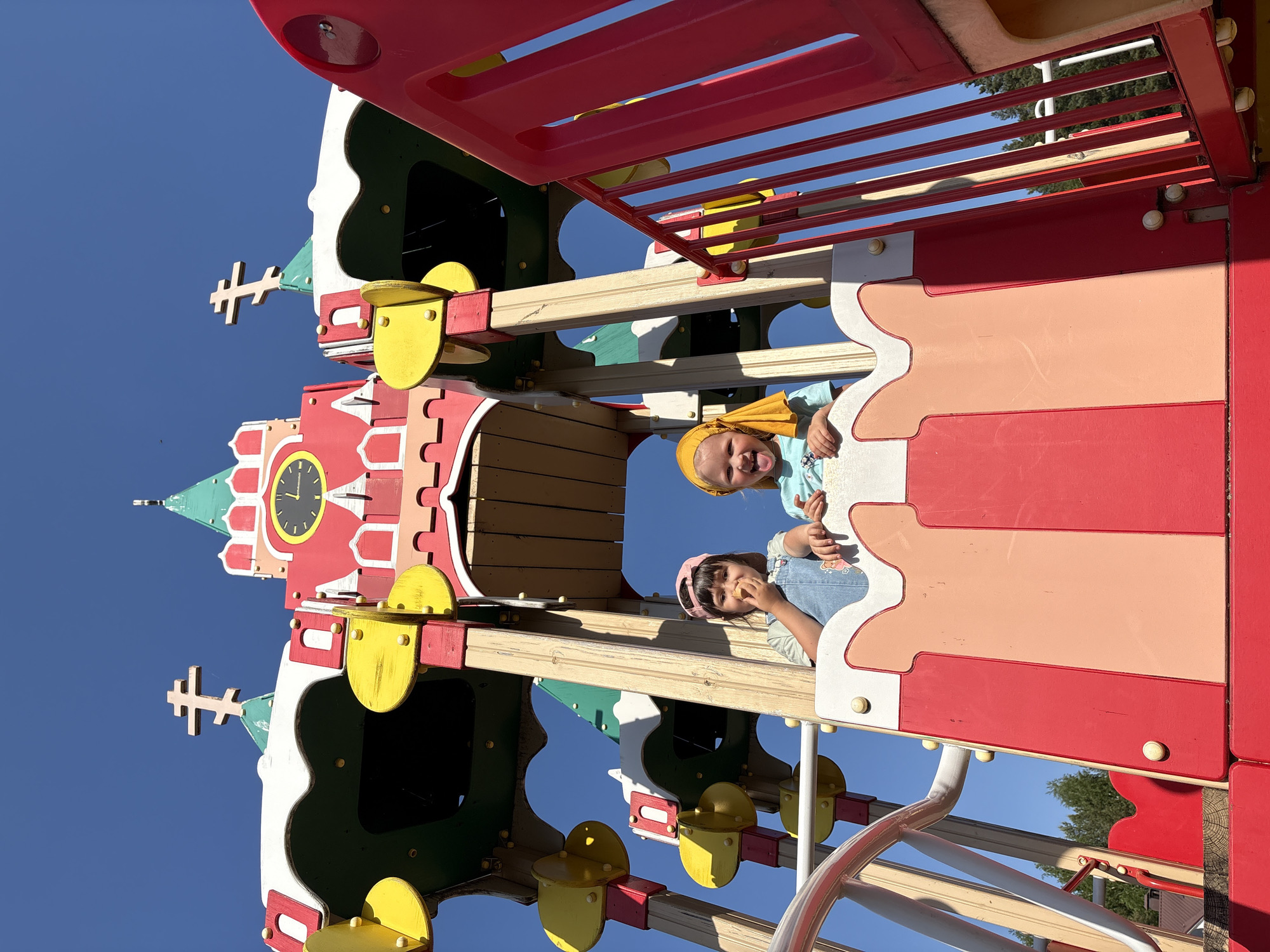 Two young girls, one wearing a yellow headscarf and the other a pink headband, peer out from a colorful playground structure. The structure, possibly located in the city of Chaikovsky, Russia, based on the architectural details of the playground equipment, features a clock tower design with red, yellow, and white elements. The background shows a clear blue sky. The girls appear to be interacting with each other, possibly playing or posing for a photograph. The playground also has additional features such as bars, platforms, and slides.