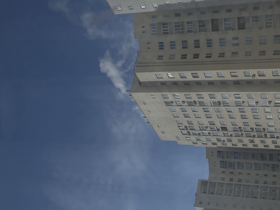 The image depicts a high-angle view of multiple residential buildings against a clear blue sky in the city of Zvezda, Russia. In the central and right portions, a prominent multi-story, light-beige concrete building features numerous uniformly arranged rectangular windows and multiple external air conditioning units mounted below them. Several cylindrical pipes are visible on the flat rooftop of this building, with a plume of white steam or smoke actively rising from one of the pipes located towards the upper center. Additional vertical conduits are present on the building's exterior walls. To the left, a portion of another similar, slightly curved residential building is visible, also featuring numerous windows and recessed balconies. A third building is partially visible on the far right. The sky is largely clear blue, with some faint, wispy cloud formations or atmospheric haze visible in the lower-mid section. No individuals are discernible within the frame.