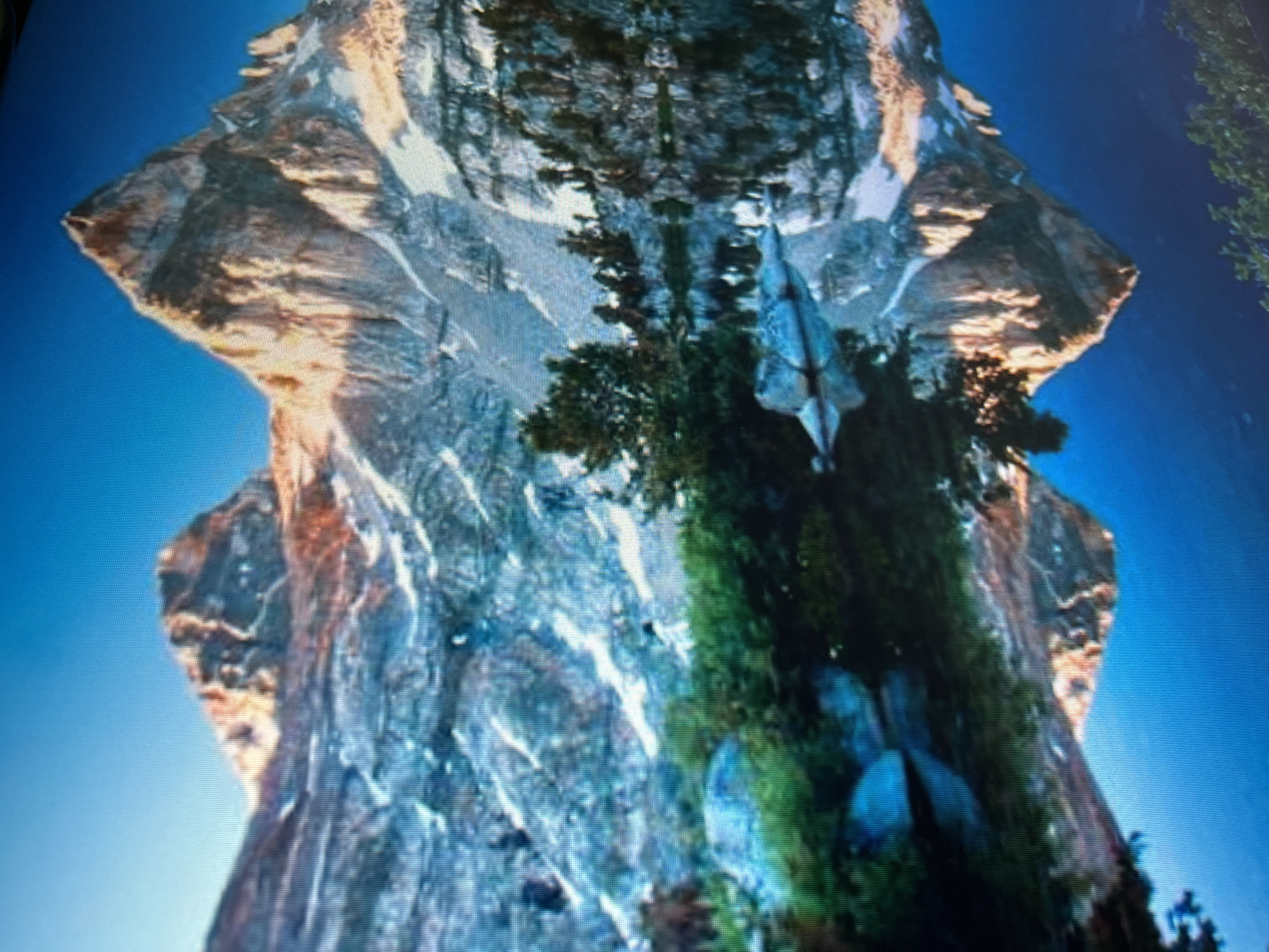 The image shows what appears to be a digitally manipulated or reflected image of a large, rocky formation with vegetation. The rock face has varied textures and coloration, suggesting it's a natural geological feature with mineral deposits. A patch of green vegetation, possibly trees and shrubs, grows on one side of the rock. The background is a clear blue sky. The composition is symmetrical, creating a mirrored effect. The specific location in Kryvyi Rih, Ukraine cannot be determined from the image provided. There are no people visible.