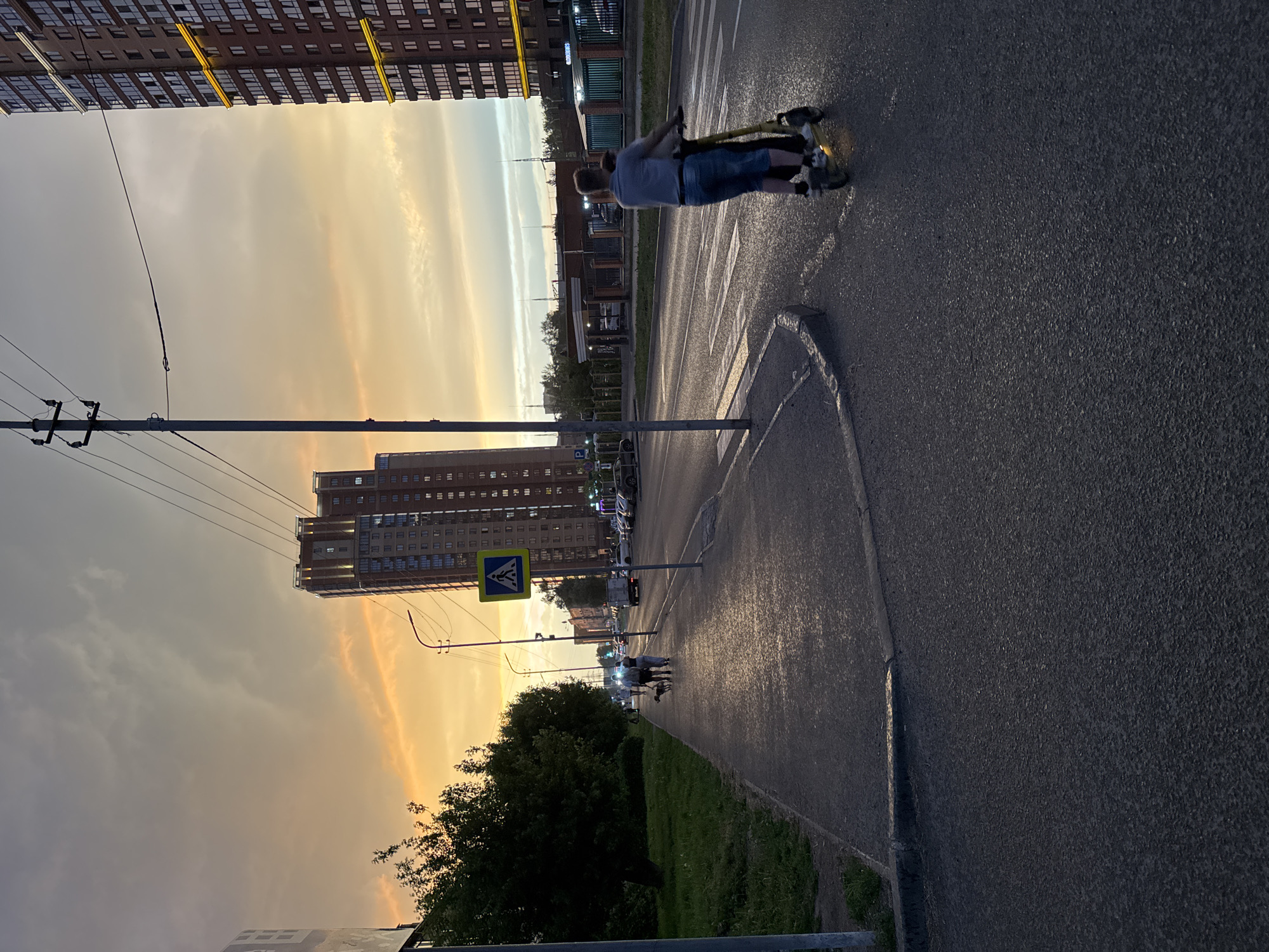 The image captures a sunset view of a street in Novosibirsk, Russia. A man is riding an electric scooter on the right side of the road, while on the left sidewalk, two people are walking a dog. Tall, modern apartment buildings with illuminated windows are visible in the background, and overhead power lines stretch across the sky, which is painted with hues of orange and gray clouds. The road appears wet, suggesting recent rainfall. A pedestrian crossing sign is also visible on the left side of the road.