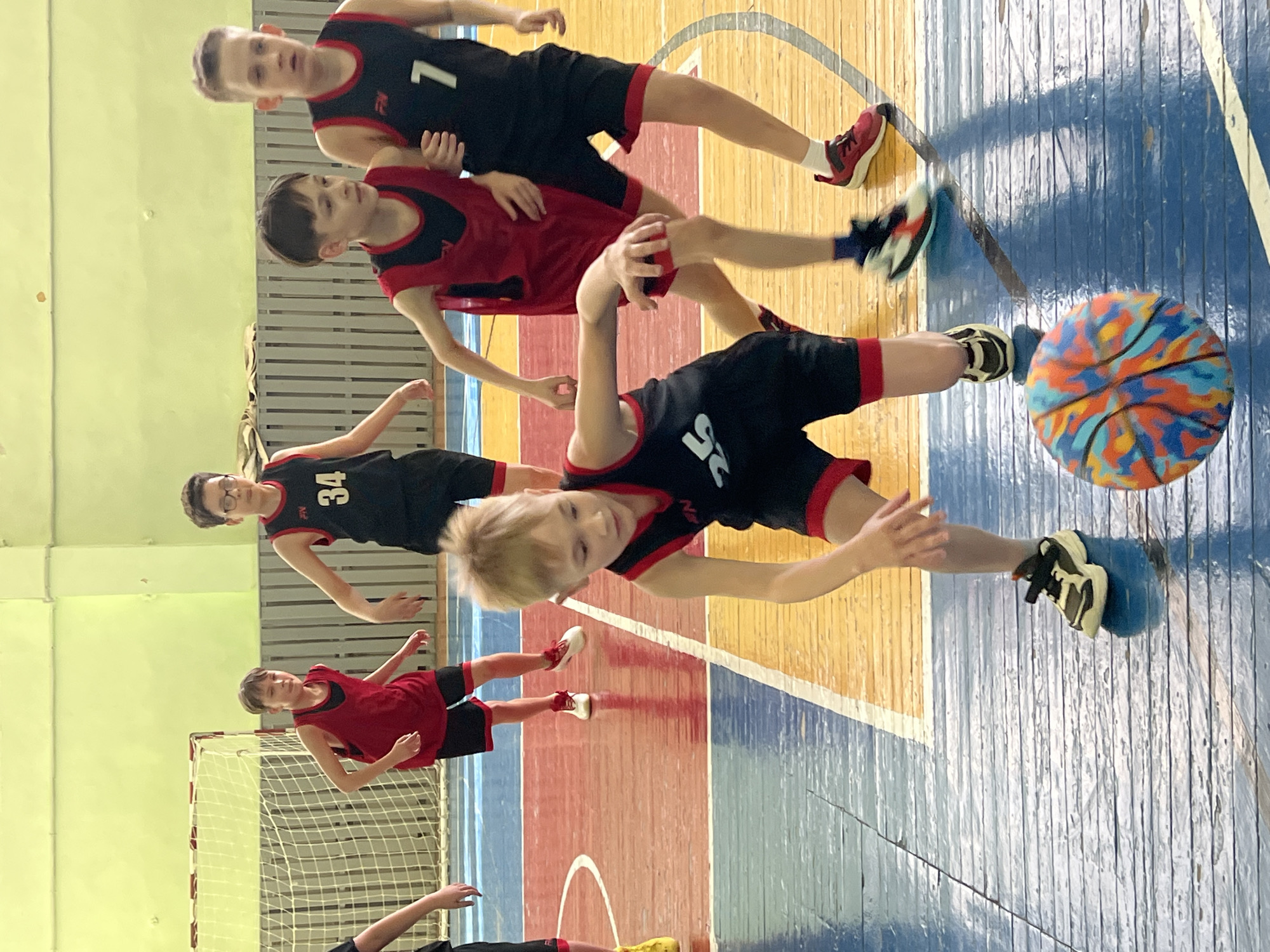 The image depicts a group of young basketball players in a gymnasium in Chaikovsky, Russia. There are five identifiable players in the scene. They are wearing athletic clothing in black and red. One player is closer to the camera, wearing a black jersey with a red trim and the number 26. He appears to be in motion, potentially dribbling or passing a multi-colored basketball. Other players are in the background, some wearing black jerseys with numbers, and others wearing red jerseys. The gymnasium has a wooden floor with painted lines in yellow, blue, and red, indicating the basketball court. A net is visible in the background on the left, suggesting a goal or boundary marker. The overall setting is a typical indoor sports facility.