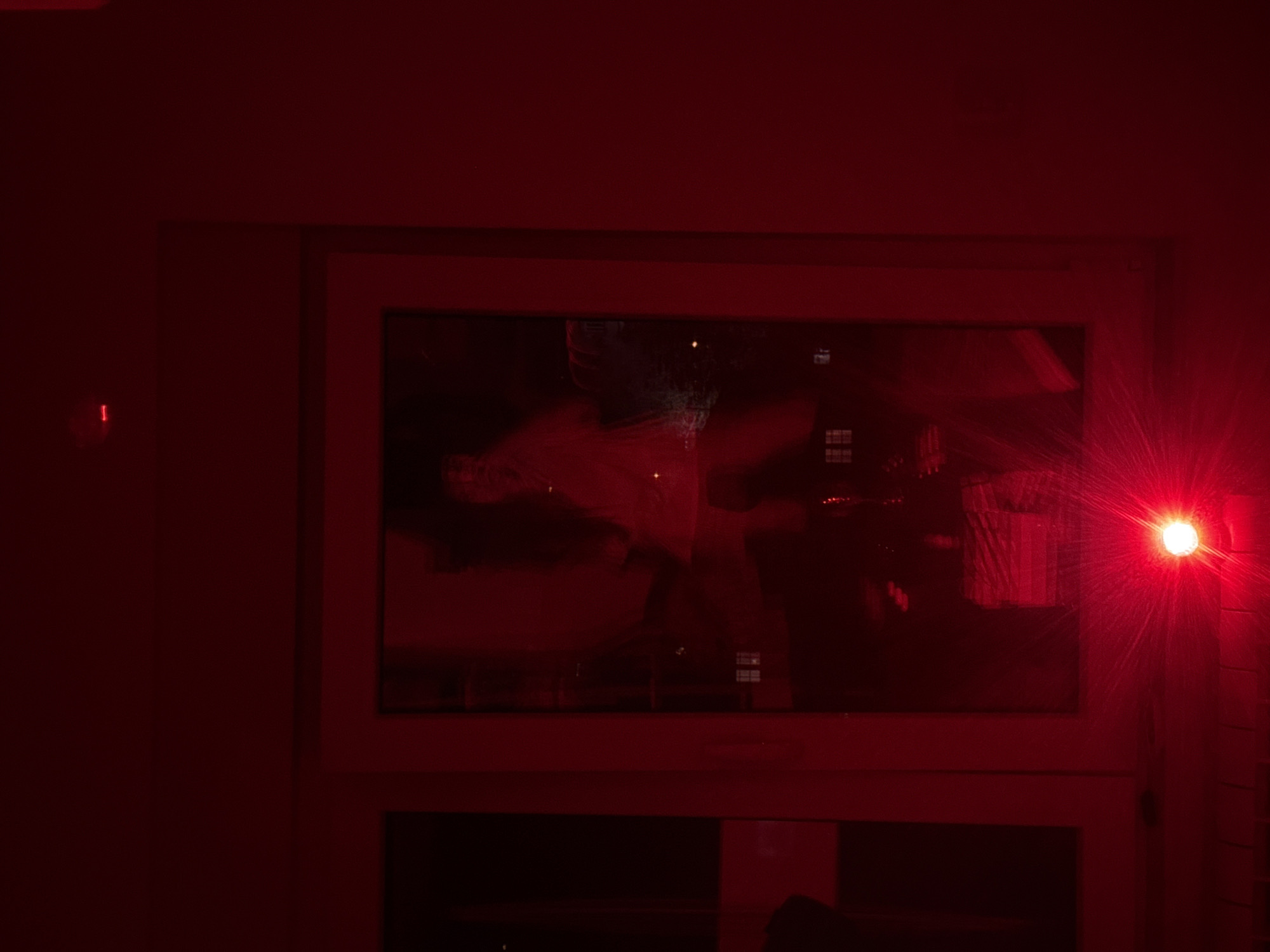 The image displays an indoor scene bathed in a strong red light, suggesting it was captured at night or in a darkened room. A prominent window, featuring a light-colored frame (rendered red by the illumination) and divided into multiple sections, occupies the central and right portions of the frame.
A very bright, starburst-like red light source is positioned on the right side, close to the window frame, acting as the primary illuminant for the entire scene. Through the window, or as reflections upon its surface, indistinct forms are visible, including what might be furniture or room elements, and possibly the hazy outline of a person. These dark shapes are silhouetted against the pervasive red light.
On the far left, a small, vertical red indicator light is discernible on a plain wall, which, like the rest of the surrounding surfaces, appears uniformly dark red. Below the main window, another horizontal section of a window or structural element is visible. No specific activity is clearly depicted, and no text is visible within the frame. The intense red lighting creates a distinctive, almost dramatic atmosphere. This scene is located in Vozdovac Urban Municipality, Serbia.