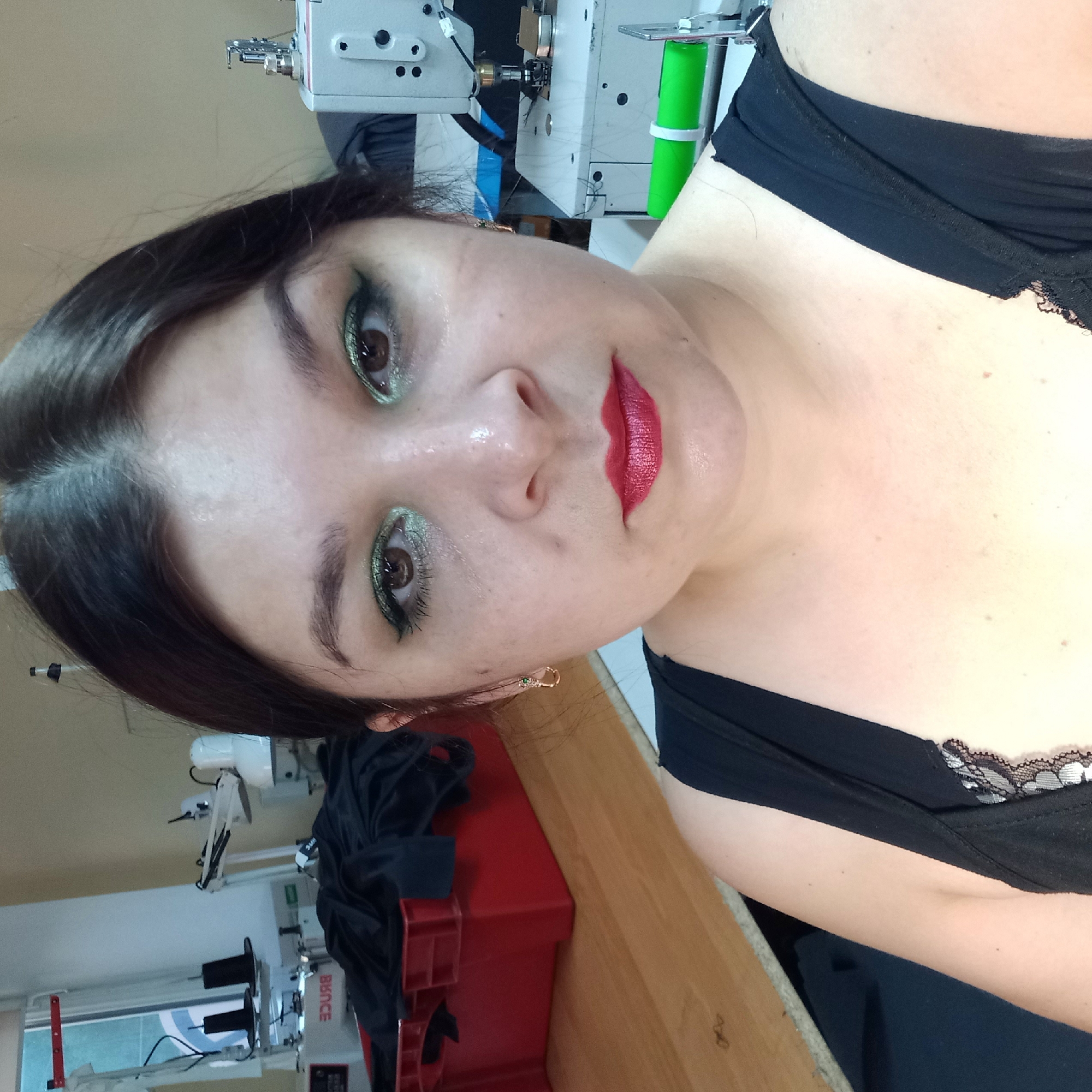 A woman with dark hair and striking green eyeshadow is taking a selfie in what appears to be a workshop or studio. She is wearing a black top with lace trim. In the background, sewing machines and fabric are visible, suggesting a sewing or garment production environment. The lighting is somewhat harsh, creating reflections on her skin. The overall impression is that of a candid moment captured in a work setting. The image does not contain any visual information to determine its specific location within Kropyvnytskyi, Ukraine.