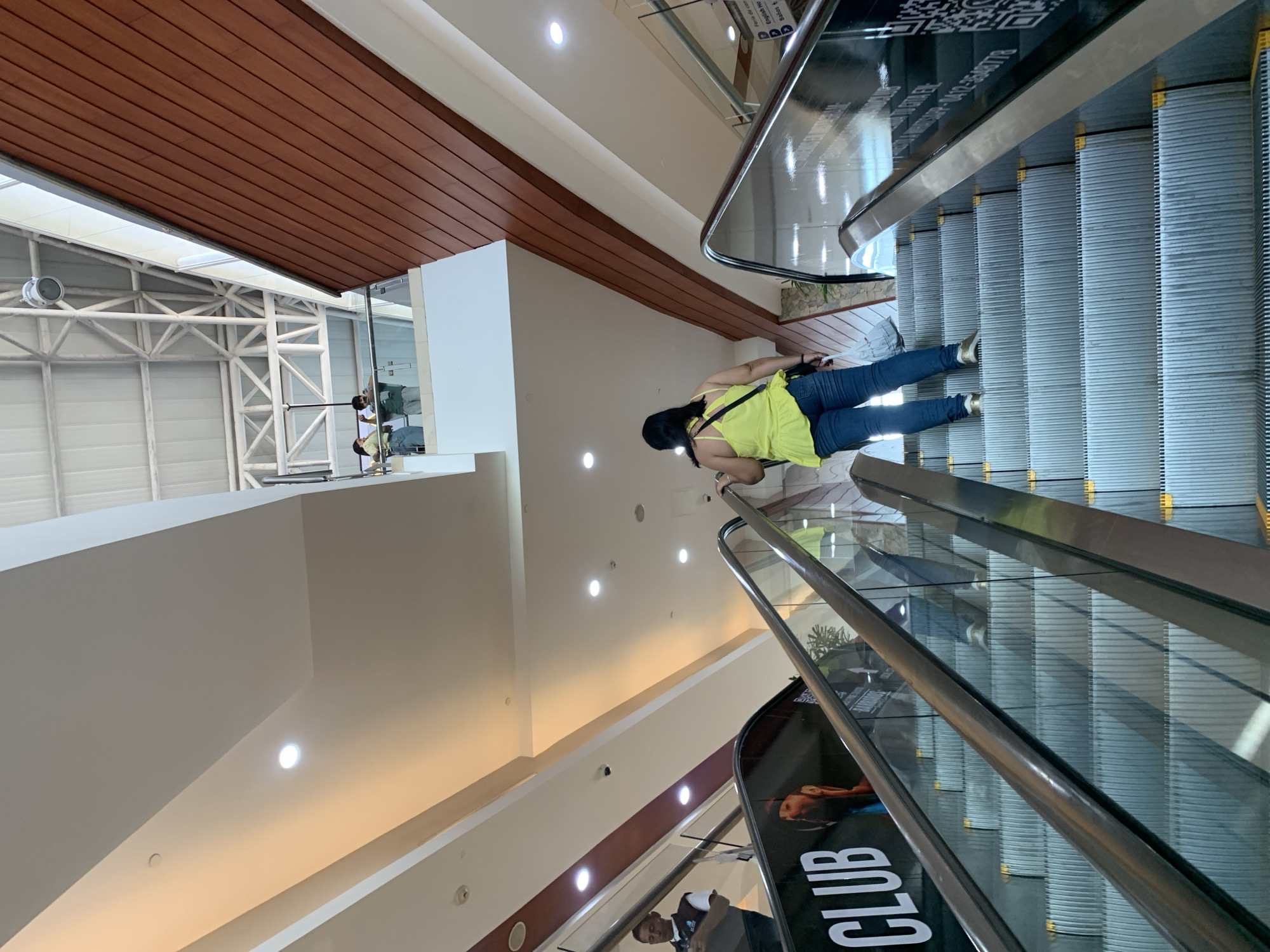 The image depicts an interior scene at the Sambil Margarita shopping mall in Porlamar, Venezuela. A woman wearing a yellow top and blue jeans rides an escalator upwards. The escalator has metal handrails and glass side panels. A promotional display with the word "CLUB" is visible on the escalator's side panel. The mall's interior features a white ceiling with recessed lighting and a section of brown wood paneling. Structural support beams are visible near the ceiling.