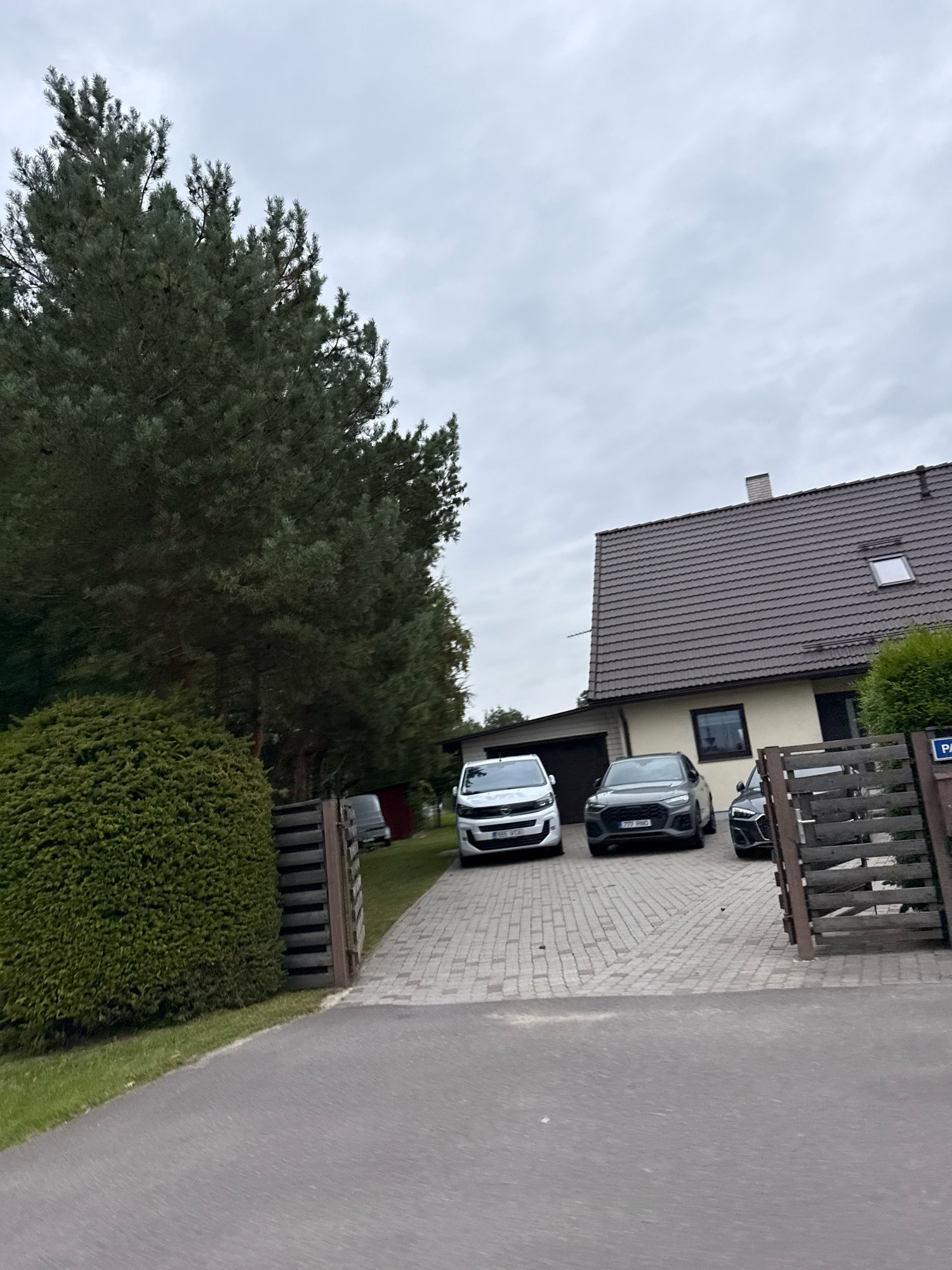 The image depicts a cloudy day scene in Peetri alevik, Estonia, focusing on a residential property. A paved driveway leads to a light-colored house with a dark tiled roof and a dormer window. Three cars are parked on the driveway: a white van on the left, a dark grey SUV in the middle, and a black sedan on the right. Adjacent to the driveway is a tall evergreen tree and a neatly trimmed green hedge. A wooden fence, partially open, stands to the right of the driveway, and a sign with the letters "PA" is visible on it. The foreground shows a grey asphalt road.