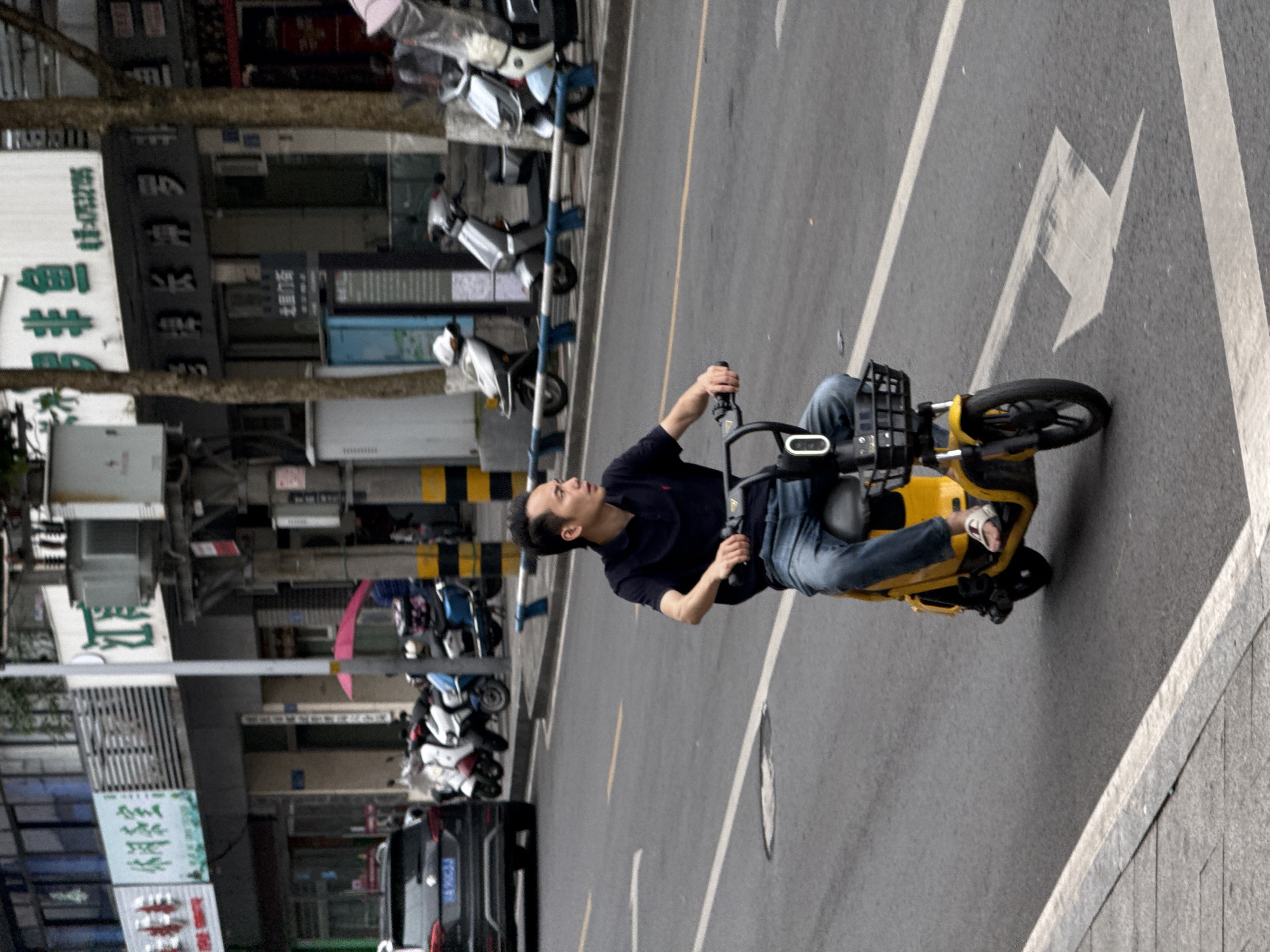 A man rides a yellow electric scooter on a gray paved road. He is wearing a black polo shirt and blue jeans, and he appears to be looking to his left. The road has white lane markings and an arrow pointing left. To the left of the road, there are parked motorcycles and scooters, as well as a black car. Buildings with Chinese signage are visible in the background. This is a street scene in the city of Jiangjin, China.