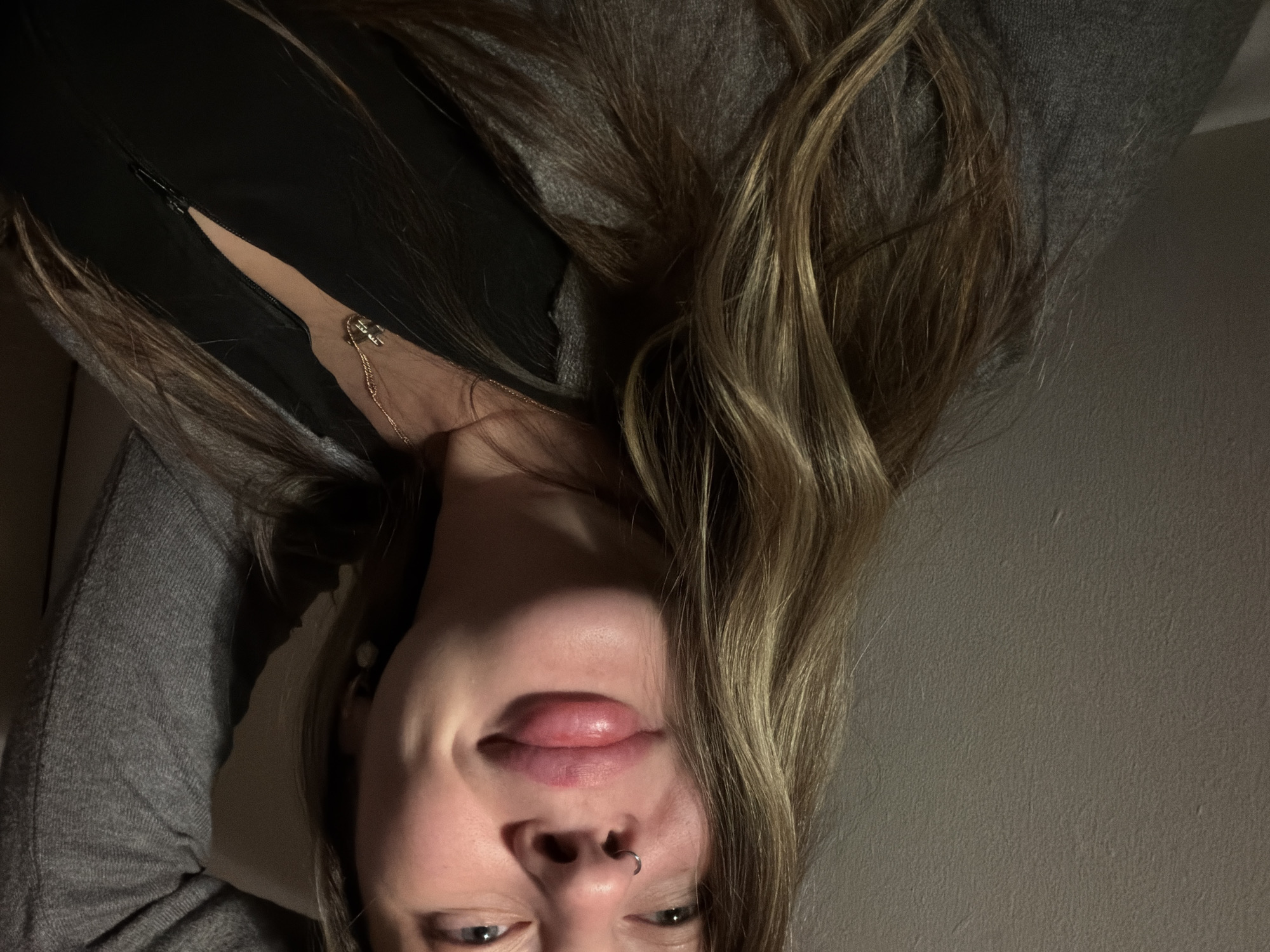 A close-up, inverted portrait features a young person with long, wavy blonde and light brown hair. The subject is wearing a grey knitted sweater or top, layered over a black garment that appears to have a zipper partially visible near the collarbone. A delicate gold chain necklace with a small, circular charm hangs around their neck. The subject has a septum piercing (nose ring) and their lips appear full, with a slight, playful smirk on their face. Their eyes are looking towards the camera, upward from their inverted position. The background is a plain, light grey wall, and the lighting is dim, suggesting an indoor setting or low light conditions. No text is visible in the image.