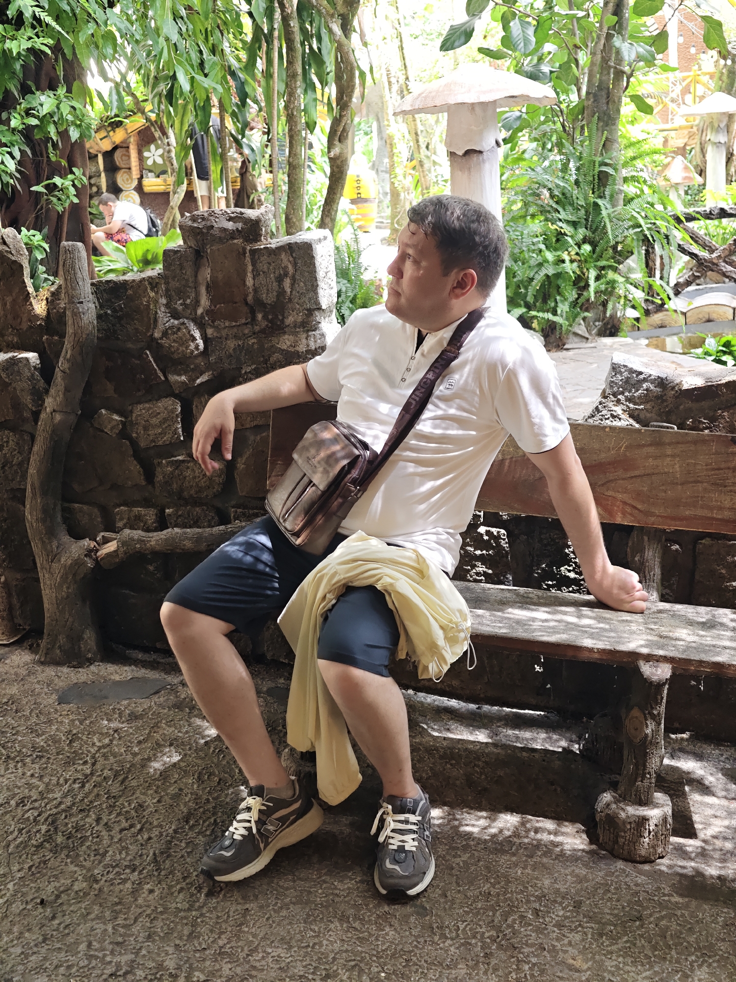 A man is sitting on a rustic wooden bench in what appears to be a garden or park setting. He is wearing a white short-sleeved shirt, dark blue shorts, and grey athletic shoes. A brown leather messenger bag is slung across his body. A pale yellow cloth is draped over his lap and the edge of the bench. He is looking to his right, away from the camera. The background features a stone wall, lush green foliage, trees, and decorative mushroom-shaped structures. The ground is a textured, earthy surface. The overall scene suggests a relaxed outdoor environment, possibly a tourist attraction in Da Lat, Vietnam, known for its gardens and unique architectural styles.