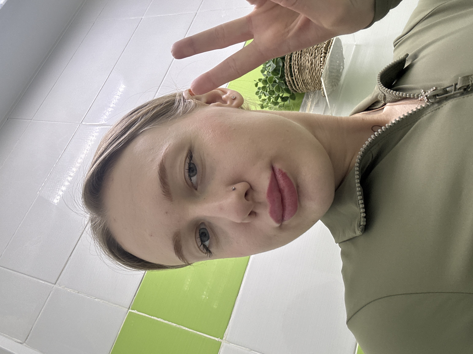 An indoor self-portrait, situated in Rostov-on-Don, Russia, depicts a light-skinned adult female, positioned from the upper chest upwards. Her head is slightly tilted, and she looks directly at the camera with her lips pursed into a pout. She wears a long-sleeved olive green collared top, partially unzipped, revealing a delicate necklace with a small, multi-faceted cross-like pendant. A small silver stud pierces her left nostril, and a small clear stud is present in her left earlobe. Her right hand, visible in the lower right quadrant of the image, displays a 'peace' sign with the index and middle fingers extended. The background features a tiled wall, with white square tiles on the upper section and lime green square tiles on the lower section. A white shelf is visible on the right, supporting a small potted plant with dark green foliage contained within a woven, light-brown basket.