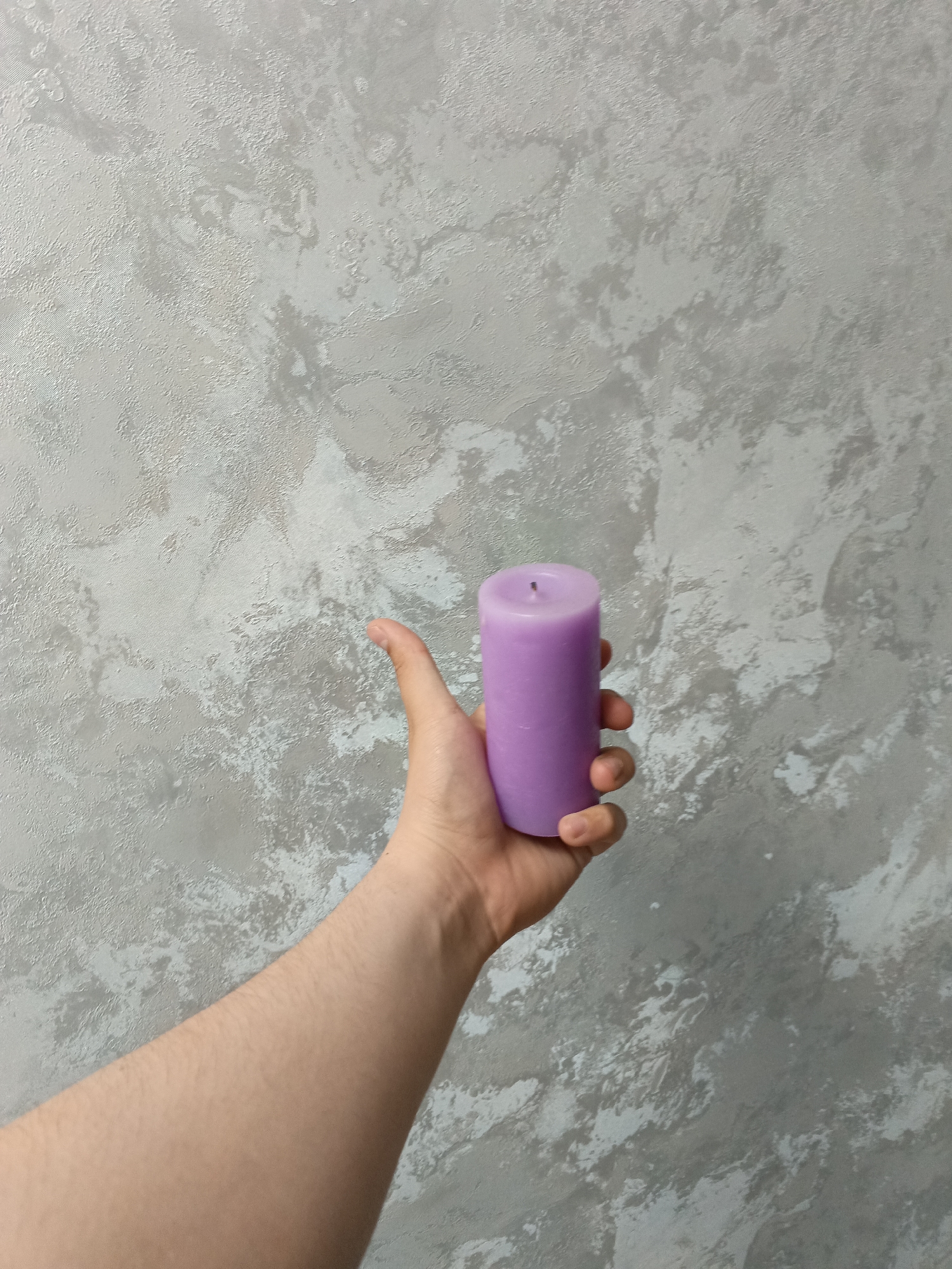 The image shows a human hand holding a purple candle against a mottled gray and white wall. The hand appears to be fair-skinned. The wall has a textured finish. There is no information available to determine a location in the city of Andreyevo, Russia.