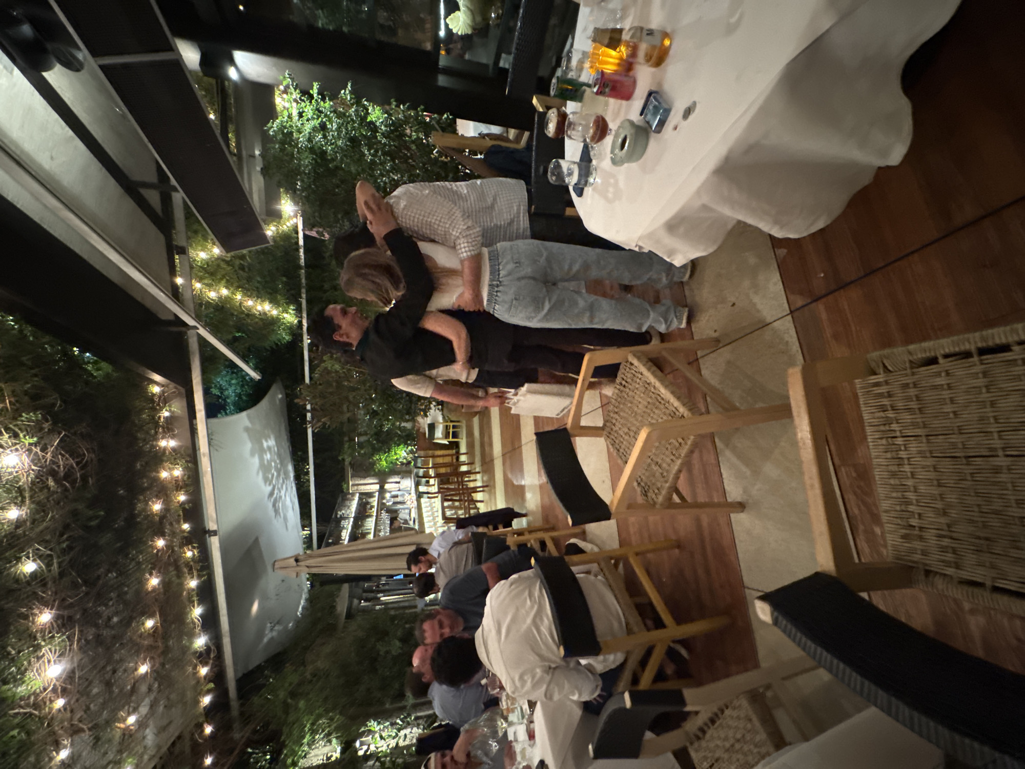 This image depicts a group of people at an outdoor dining area, likely a restaurant or bar. The setting appears to be in Mexico City, Mexico, given the vegetation and architectural style. Some individuals are standing together, with one group appearing to embrace or dance near a table with drinks. Others are seated at tables, presumably engaged in conversation or dining. String lights hang above, adding to the ambiance. The tables are covered with white tablecloths, and there are chairs and wicker furniture visible.