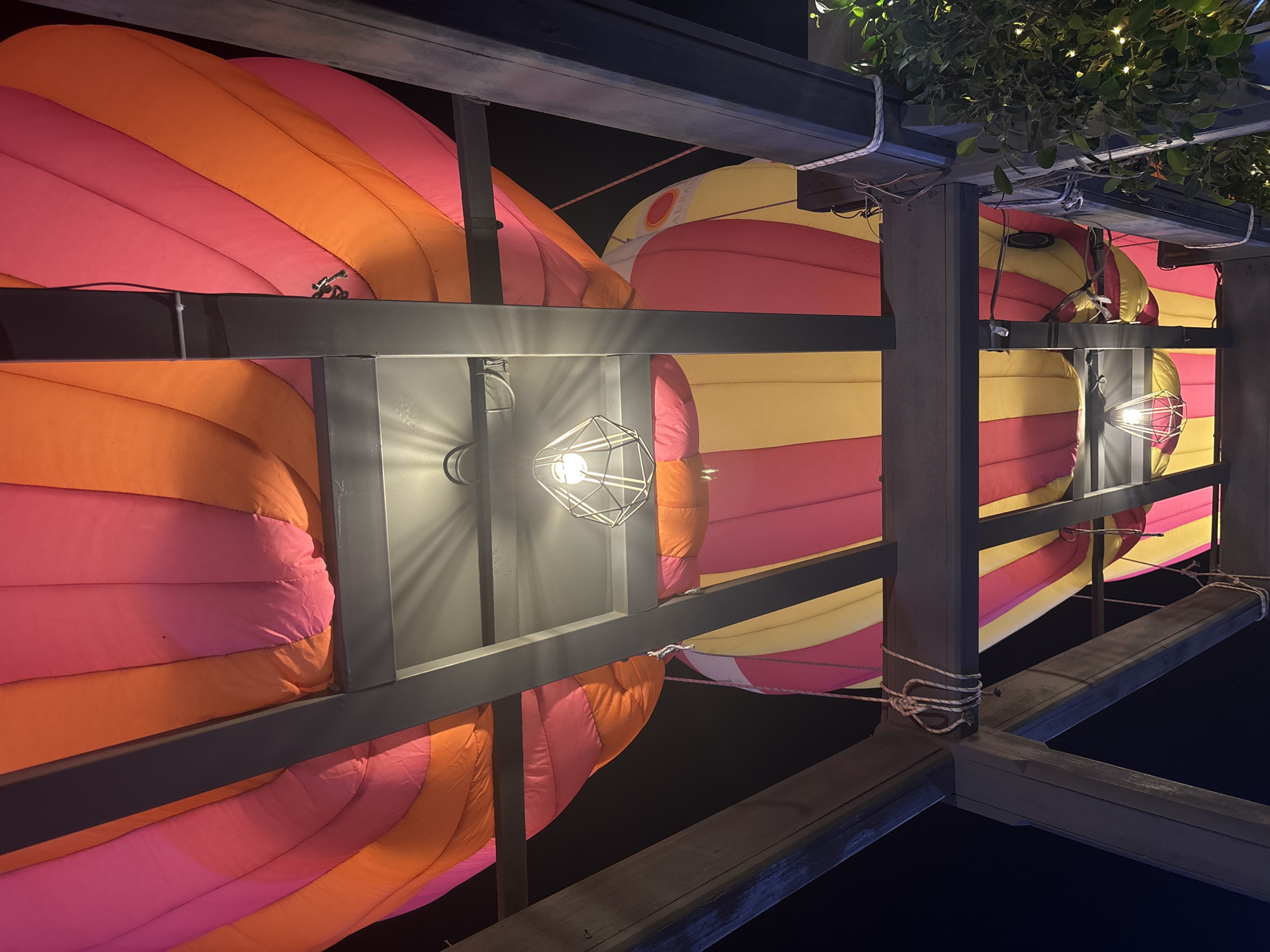 The image captures a low-angle, nighttime view of an outdoor dining area in Amman, Jordan, decorated with several large, inflatable hot air balloon replicas. These balloons are brightly colored with alternating stripes of pink, orange, and yellow. They are suspended above a wooden pergola structure, with metal beams and rafters supporting the arrangement. Two modern, geometric pendant lights hang from the structure, illuminating the scene with a warm glow and casting patterned shadows on the balloons. White ropes are used to tie down sections of the balloons and secure them to the pergola. Lush green foliage with small white fairy lights is visible on the right side of the frame, adding a touch of nature to the urban setting. The background is dark, indicating it is nighttime. No people are visible in the image.