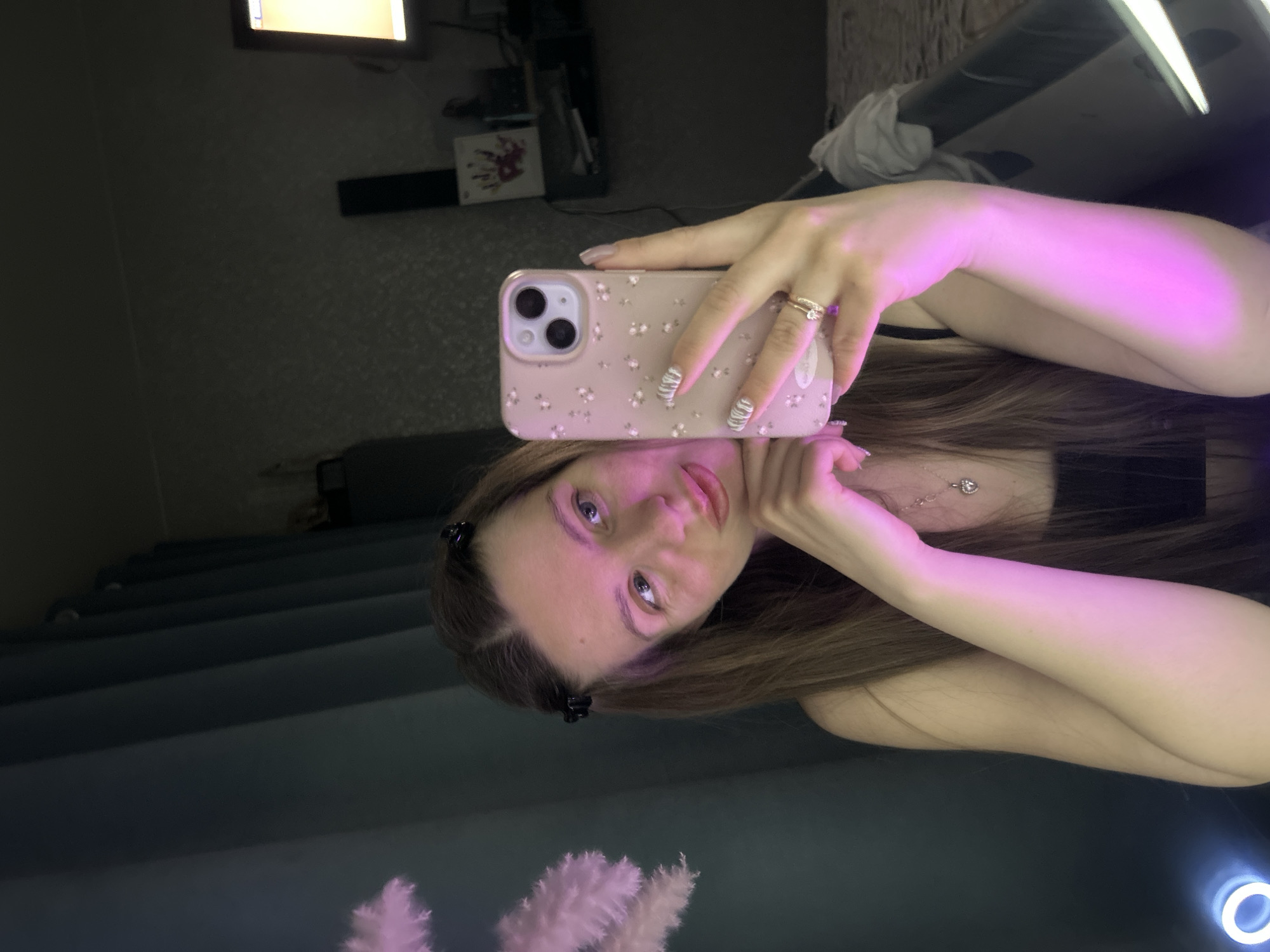 The image is a selfie taken in a room with mixed lighting. A young woman is holding up a pink phone with a floral case in her right hand, partially obscuring her face. She has long brown hair, fair skin, and is wearing a necklace. The background includes a glimpse of a room with furniture and decorations, as well as a reflection in a mirror. There is no evidence to suggest this photo was taken in Klin, Russia.