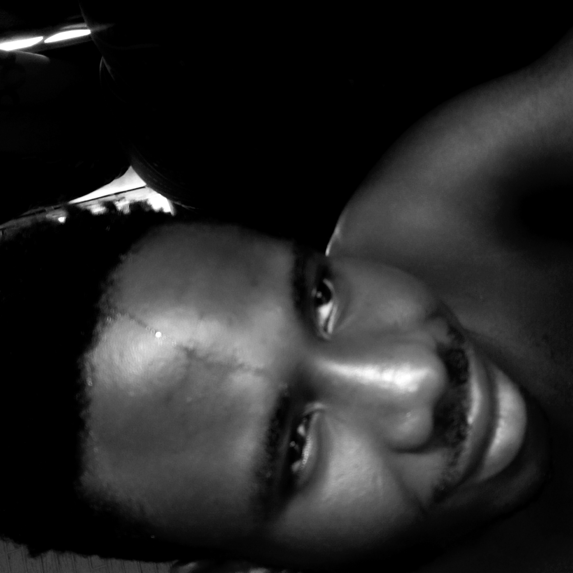 A close-up black and white image shows a man's face with a mustache. A scar is visible on his forehead. The man is lying down, with his shoulder visible in the upper right. The background is dark and indistinct. No location in Narayi, Nigeria, is discernible from the image.