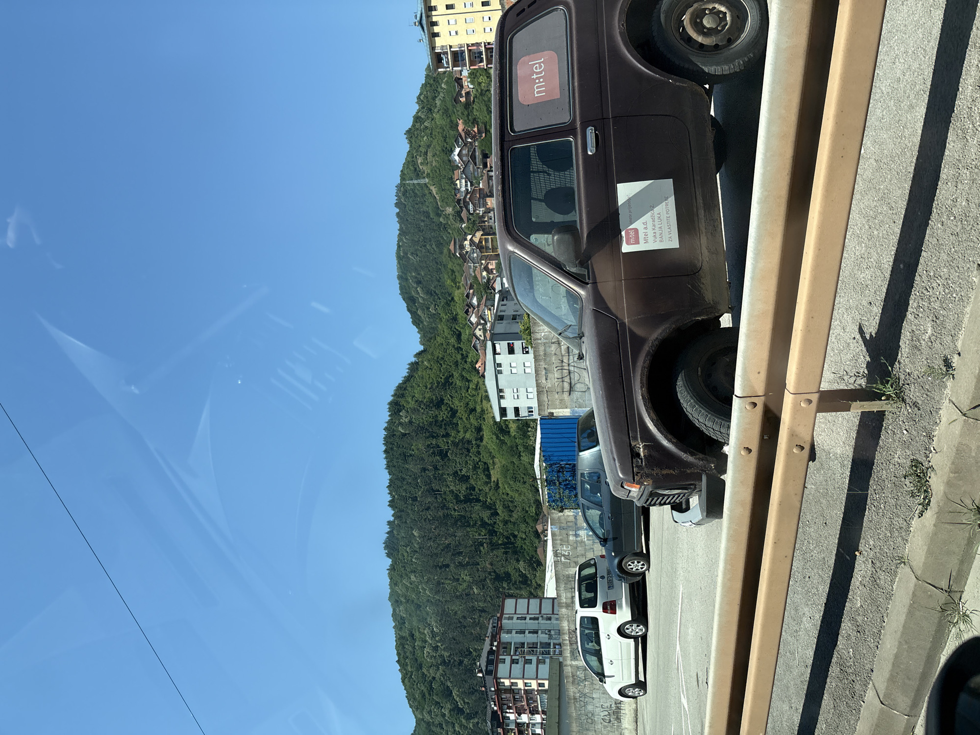 The image depicts an outdoor scene, likely in Zvornik, Bosnia and Herzegovina, with a clear blue sky and a verdant, tree-covered hillside forming the backdrop. The hillside is dotted with houses. In the foreground, a brown, older model SUV is parked on a paved surface, partially obscured by a yellow metal guardrail. The SUV has a white sticker with "mtel" in red letters on its rear passenger window. Another white rectangular sticker with text in a foreign language and "mtel a.d." is visible on the side of the vehicle. To the left of the SUV, a dark gray sedan and a white minivan are also parked. Behind the vehicles, a concrete wall with graffiti is present, and beyond that, several multi-story buildings are visible. The overall impression is of a daytime scene in a semi-urban or suburban environment.