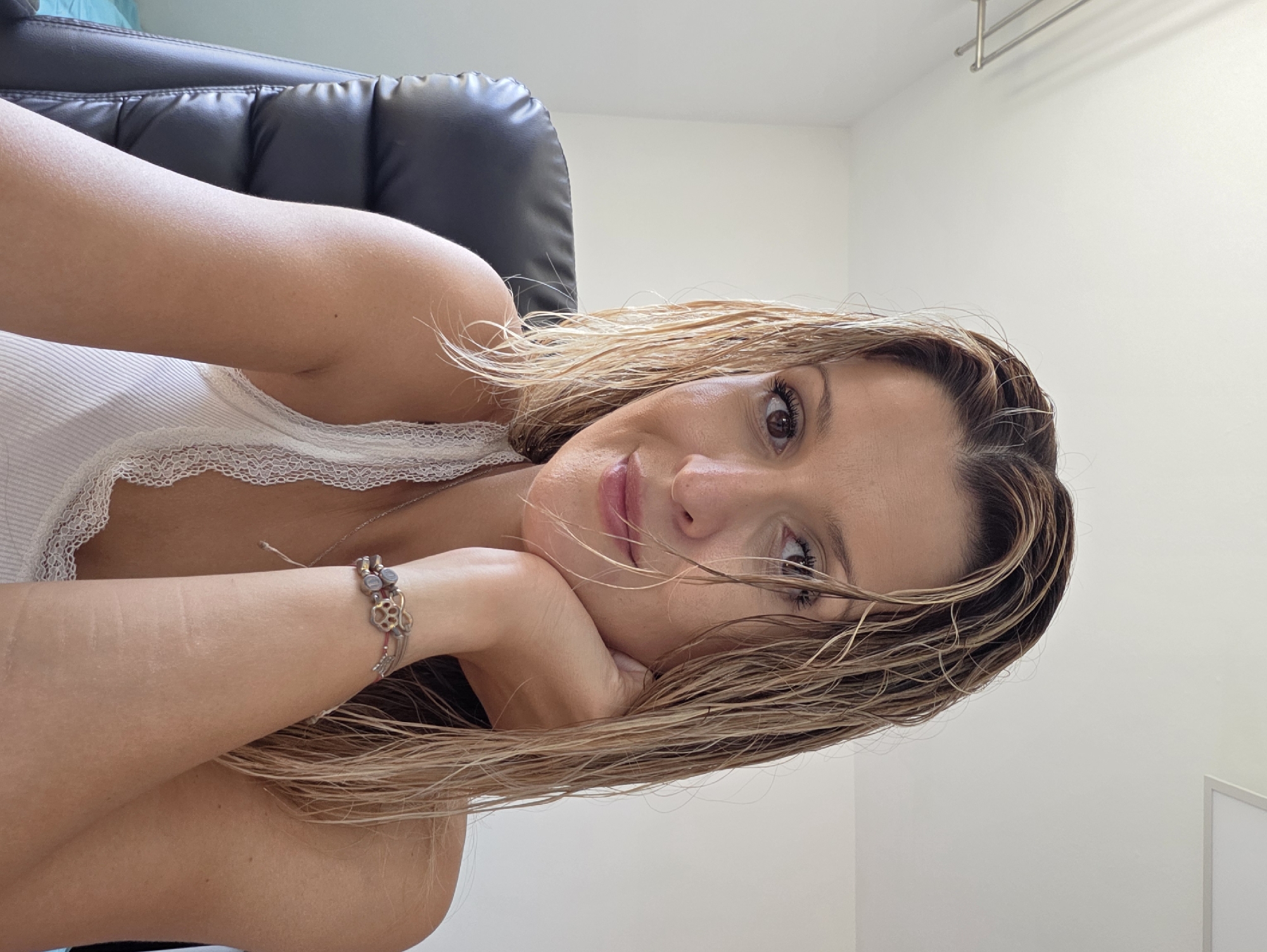 A woman with wet, blonde hair rests her chin on her hand as she looks at the camera. She is wearing a white, ribbed tank top with lace trim. A bracelet with charms adorns her wrist. She is seated in a black office chair in front of a white wall. The scene appears to be indoors, possibly an office or home. There is no indication of the location being in Munich, Germany.