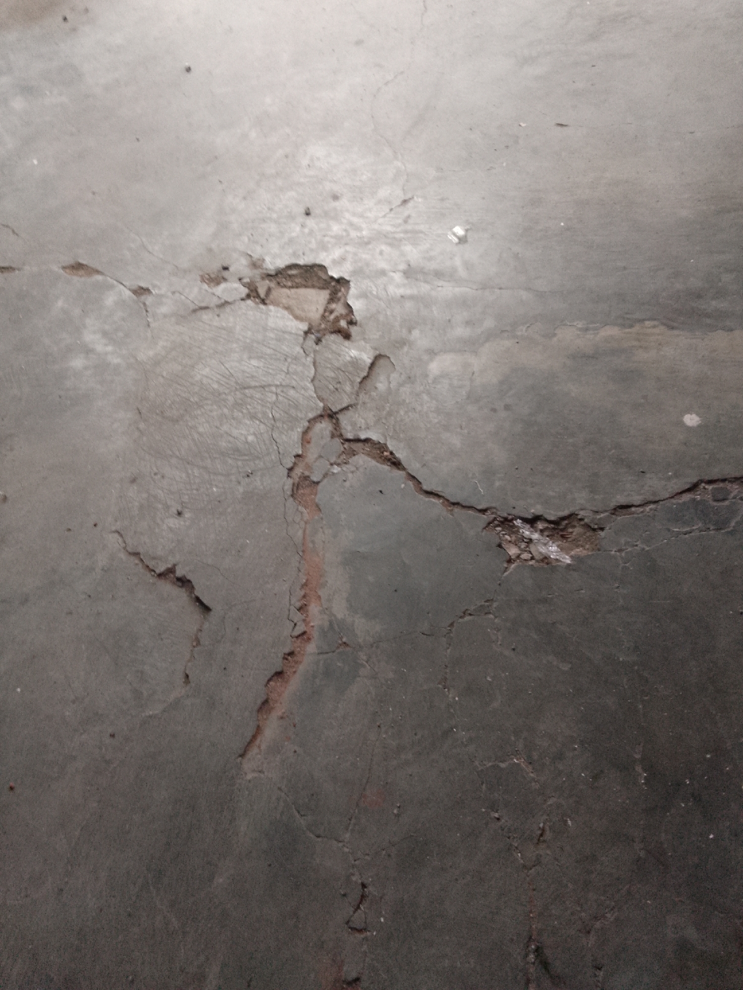 The image displays a cracked and damaged concrete floor. The cracks are irregular and vary in width, with some areas showing exposed aggregate or reddish-brown soil beneath the concrete surface. The concrete itself appears weathered and shows signs of wear, possibly from foot traffic or impact. The lighting suggests an indoor environment, possibly a basement or utility area, with a dull, gray ambiance. The scene is devoid of any human presence or other distinguishable objects. The condition of the floor indicates a need for repair or maintenance. Given the image alone, it is impossible to determine its specific location within Trenggalek, Indonesia.