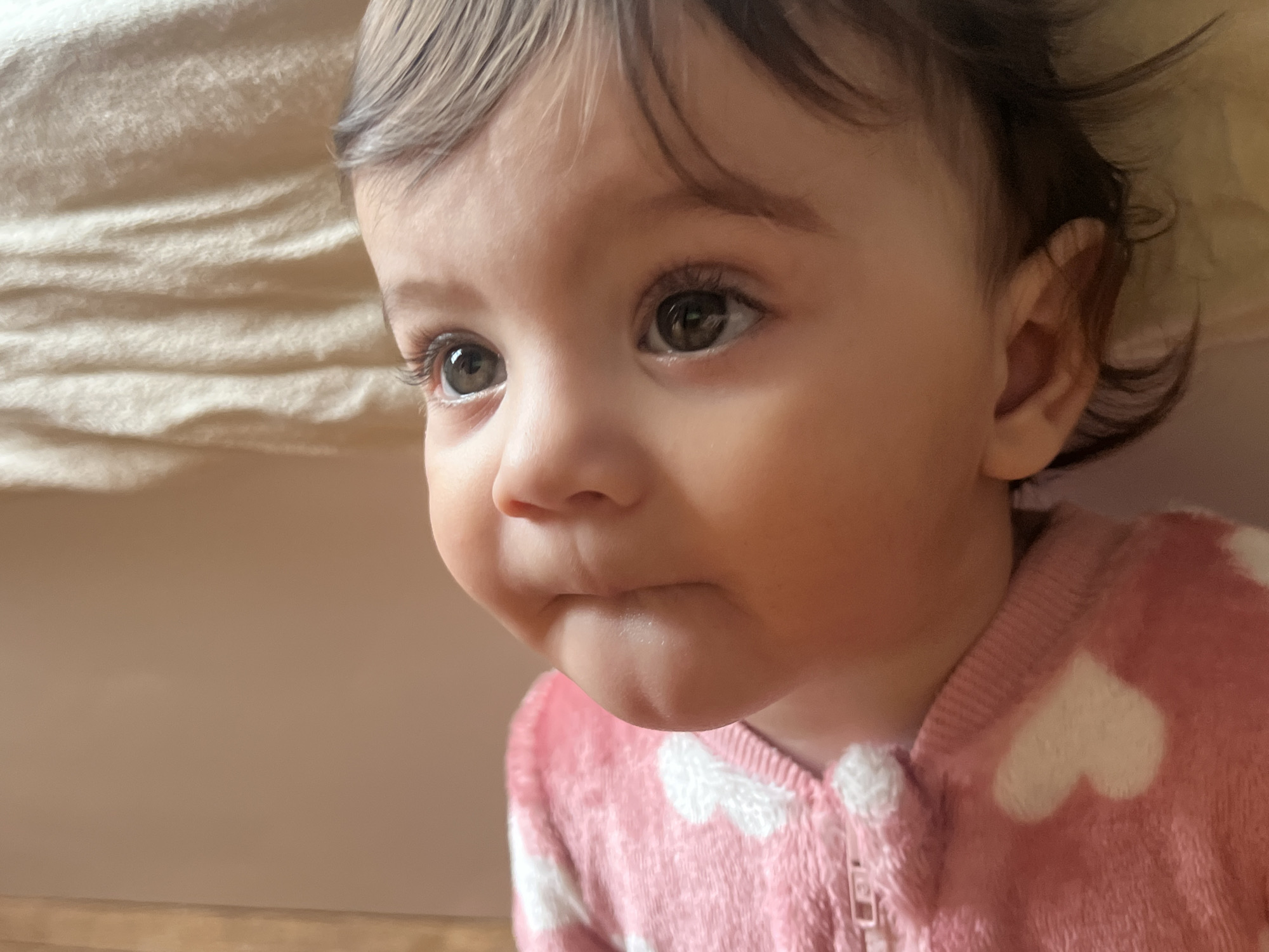 A close-up shot of a baby girl with light skin, dark hair, and brown eyes looking off to the side. She is wearing a pink onesie with white hearts. The background is a light, neutral color, possibly a wall or bedding. The location is not identifiable from the image alone; no information is available that would suggest the location is Elena, Bulgaria.
