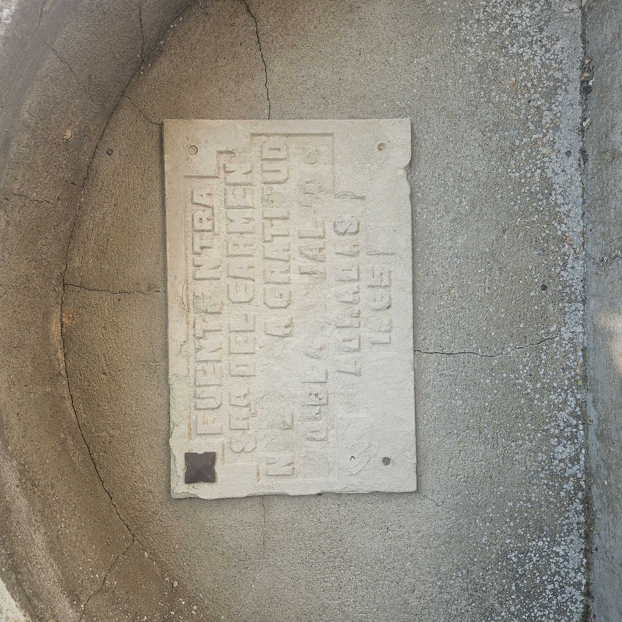 The image shows a stone plaque affixed to a concrete structure, likely a well or fountain. The plaque is inscribed with the text "FUENTE NTRA SRA DEL CARMEN EL PBLO A GRATITUD AD ALIAS TECE". A square or floral decorative element is present in the lower-left corner of the plaque. The concrete structure is aged and cracked. This is located in Algora, Spain.
