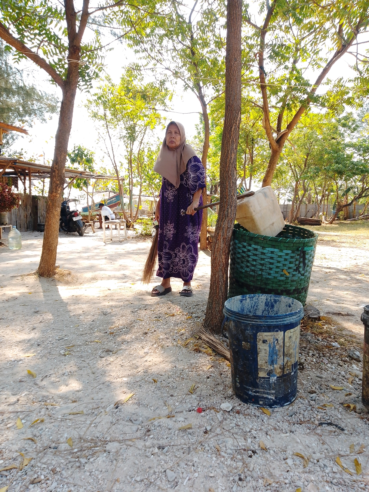 An elderly woman with a light brown headscarf and a purple floral patterned dress stands outdoors, holding a broom. She is positioned between two trees. To her right, there is a large green woven basket containing a light-colored plastic container and a blue bucket. The ground is covered in sand and small stones. In the background, a motorcycle is parked near a simple wooden structure. The scene appears to be in a rural or coastal area, possibly in Cikal, Indonesia, characterized by natural foliage and rustic elements.
