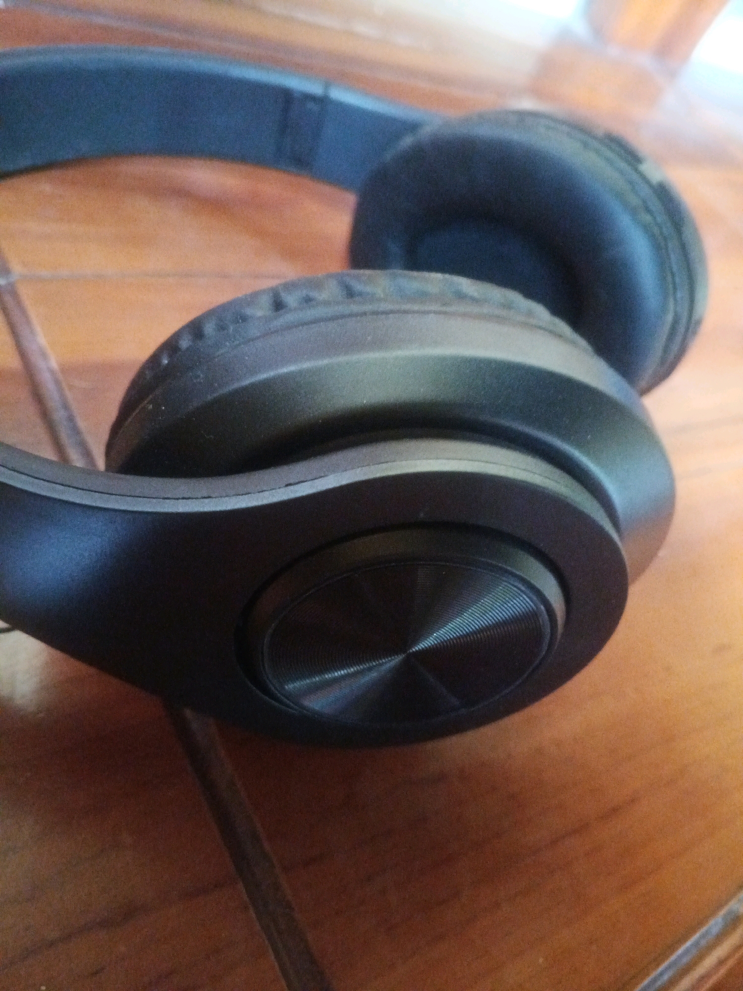 The image shows a close-up, low-angle shot of a black over-ear headphone. The headphone is resting on a wooden surface, likely a table or floor, which has a warm, reddish-brown tone and visible wood grain. The lighting appears to be natural and somewhat diffused, casting subtle shadows. The headphone itself is dark and matte, with a padded earcup and a textured detail on the outer side of the earcup, resembling concentric circles. The headband is visible extending upwards and out of the frame. No people or actions are depicted. The environment suggests an indoor setting. The image does not contain any information to suggest a location in Mewek, Indonesia.