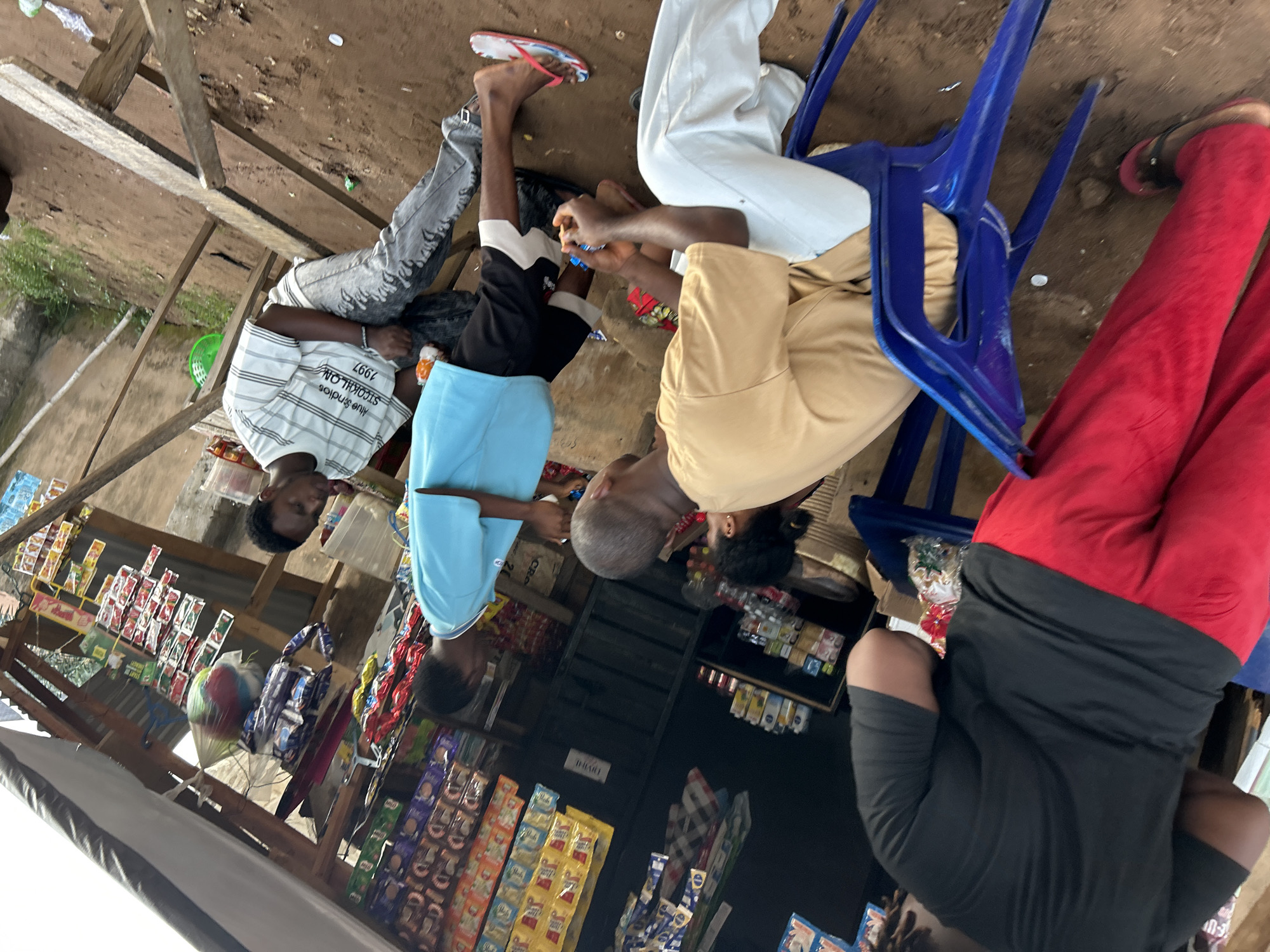 In Ihiagwa, Nigeria, an outdoor market stall is depicted with various goods displayed. Several individuals are present. In the foreground, a person wearing a black t-shirt and red trousers is partially visible. To their right, two men are seated. One man is wearing a tan t-shirt and white trousers, and the other, in a blue t-shirt and dark shorts, is standing. Further back, another man in a striped t-shirt and grey jeans is visible. The stall itself has shelves stocked with packaged goods, and an awning provides shade. The ground is dirt, typical of outdoor market environments.