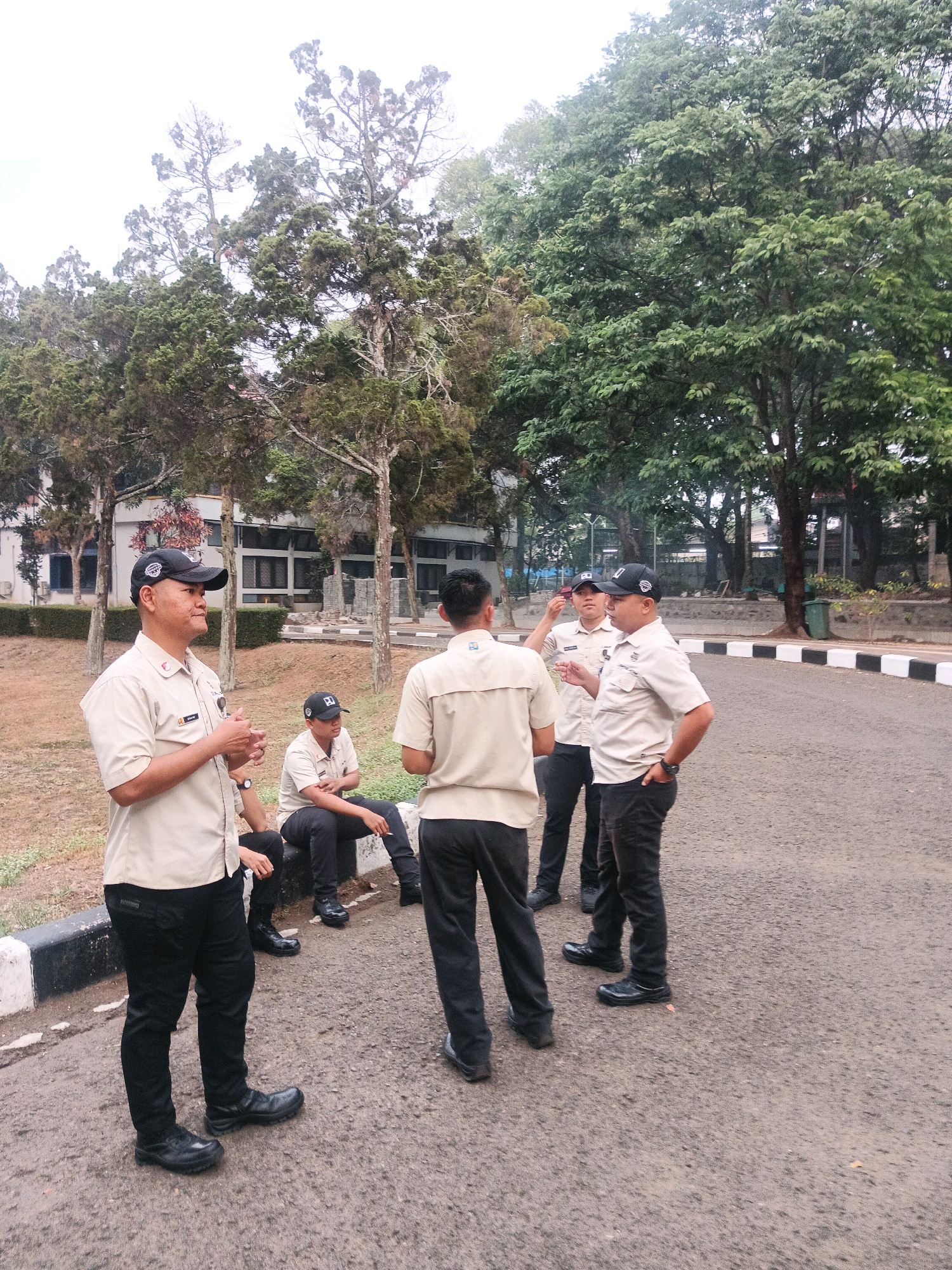 A group of five men, all wearing tan collared shirts, black pants, and black caps, are standing and sitting on a paved road in front of a building with trees surrounding the area. The man on the far left, facing mostly towards the left, is speaking and gesturing with his hands. A man in a tan shirt and black pants sits on a dark object to his right. Another man, also in a tan shirt and black pants, sits behind him. Two men in similar attire stand to the right, facing each other and conversing. The building in the background appears to be an office or institutional structure. The road has white and black painted curbs. The environment suggests an outdoor setting, possibly a campus or a corporate complex. The presence of uniforms suggests they may be security personnel or employees of a company. The overall atmosphere is casual, with the men engaged in conversation. The scene is set during daylight hours, with overcast skies. The image quality is moderate, with some graininess. The location is likely in the city of Bandung City, Indonesia, given the context of the request.