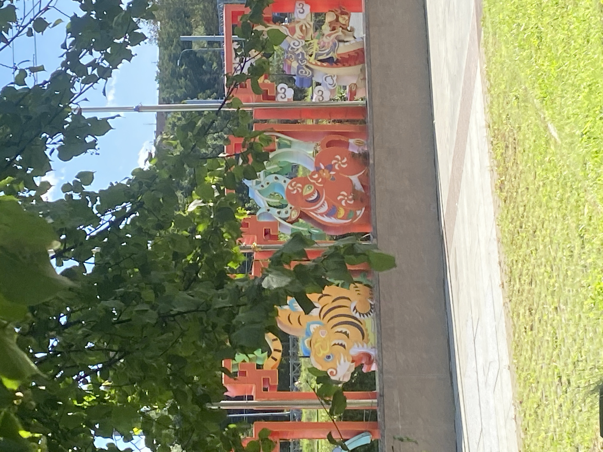 A close-up, slightly blurry shot captures a row of colorful decorative panels positioned behind a low concrete wall. The panels feature stylized depictions of animals and figures, with prominent orange and yellow elements. A tiger, rendered in yellow and black stripes with blue eyes, is visible on the left. To its right, a rounded orange figure with swirling white patterns and green accents is present. Further to the right, a figure in white and red, possibly a traditional opera character, is depicted with intricate headdress details. The background includes green foliage from a tree in the foreground, a bright blue sky with scattered clouds, and some distant trees and urban structures. The scene is set outdoors, likely in a park or public space within Saint Petersburg, Russia, as indicated by the presence of typical urban infrastructure and vegetation consistent with the region. The lighting suggests it is daytime.