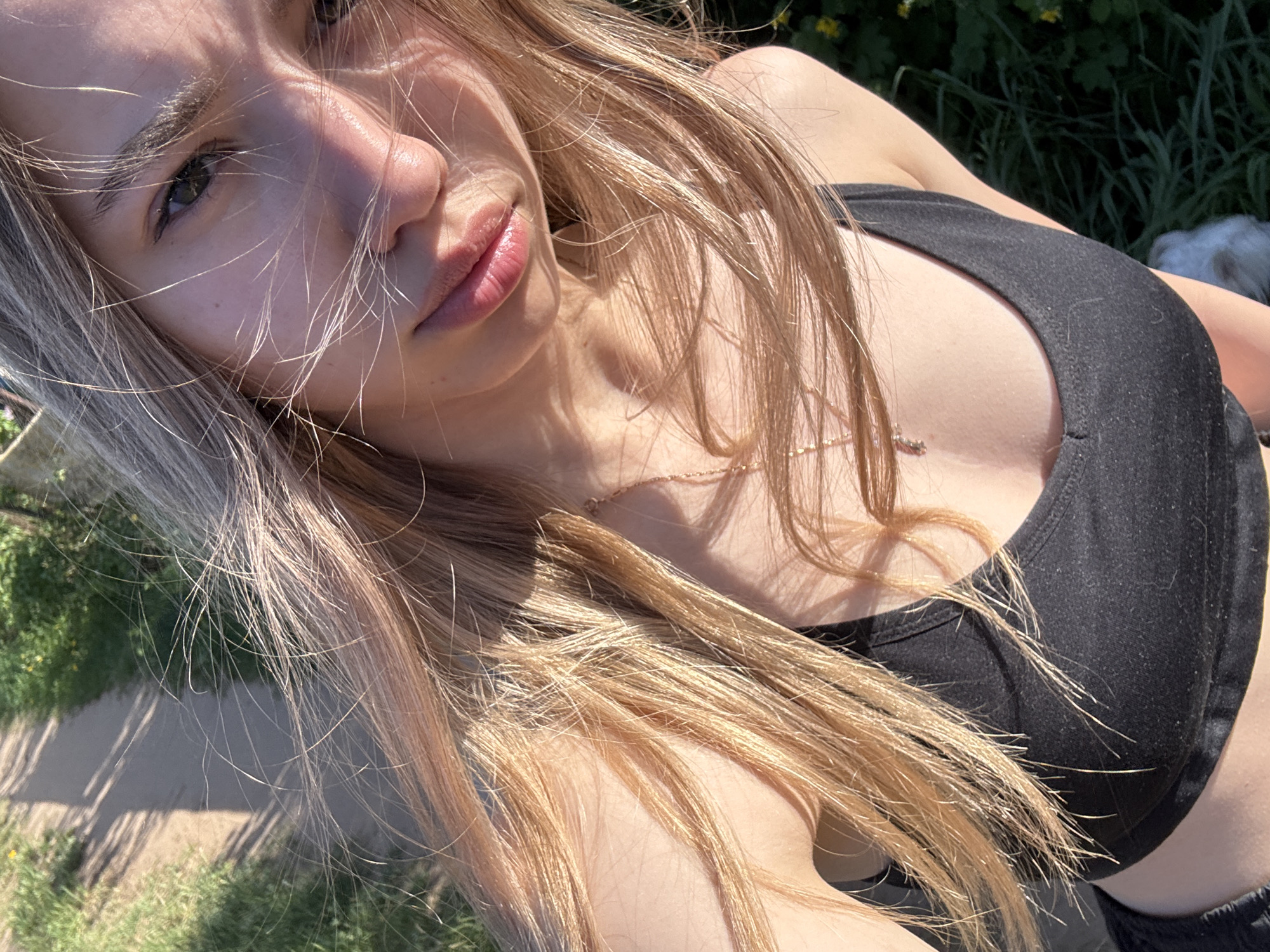 A young woman with blonde hair is outdoors in Noviy, Russia. She is wearing a black top and a gold necklace. The environment includes grass and a partial view of a sidewalk.