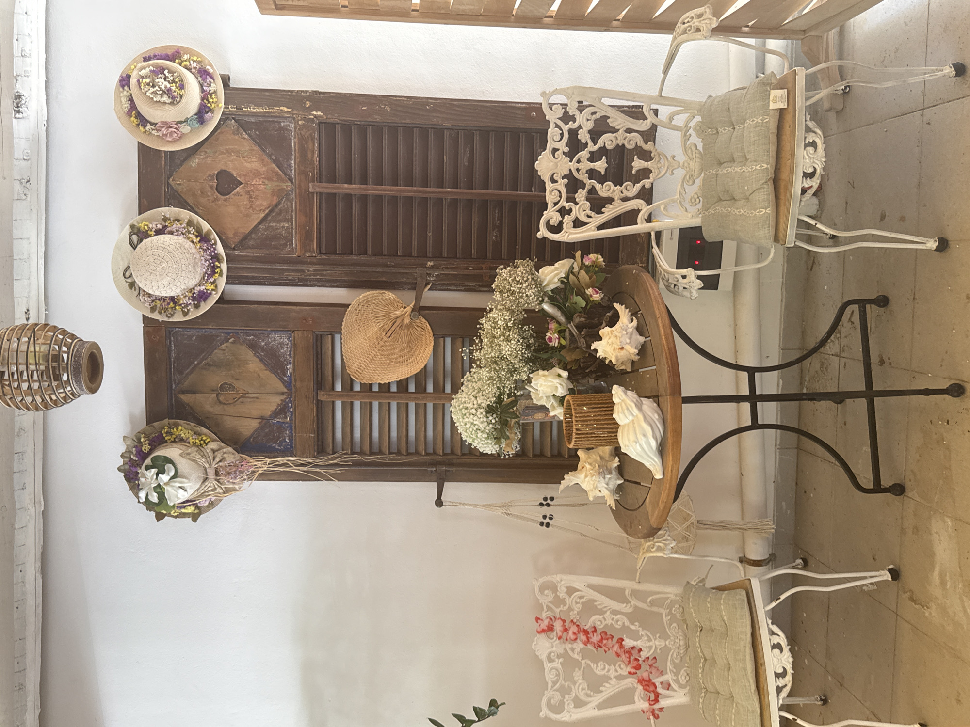 The image depicts a quaint, rustic scene that evokes a Mediterranean ambiance, likely in Rhodes, Greece, given the decorative elements. Positioned against a textured white wall is a pair of dark wooden shutters, with carved heart motifs and a diamond-shaped inlay, suggesting an aged, possibly historical setting. To the left, an ornate white wrought-iron chair with a light green cushioned seat is partially visible, adorned with a red floral garland.
On the right, a similar white wrought-iron chair with a matching cushion is also present, facing slightly away from the viewer. Between the chairs, a round wooden table with a darker border stands on a black metal base. The table is laden with decorative items, including several large seashells, a woven cylindrical container, and a bouquet of white flowers, possibly baby's breath and roses, in a glass vase.
Above the table and shutters, several decorative straw hats adorned with flowers and ribbons hang on the wall, adding to the charming, artisanal feel. A large, woven pendant light fixture is suspended from the ceiling. A hand-held fan is also displayed on the shutters. The floor appears to be tiled or concrete, with some visible stains or marks. The overall impression is one of a welcoming, outdoor seating area, perhaps a patio or garden café, decorated with a coastal and vintage aesthetic.