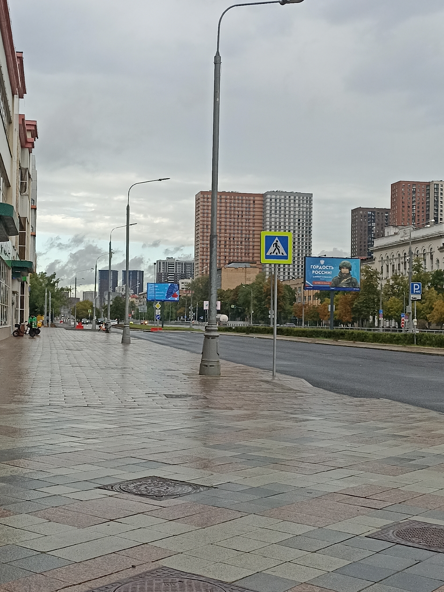 The image depicts an overcast day on a city street in Moscow, Russia. The foreground is dominated by a wet, tiled sidewalk with several manhole covers. To the left, a modern building with a white facade and red-trimmed roofline lines the street. Further down the sidewalk, two individuals on scooters, one with a green delivery bag, are parked.
The street itself is asphalt and appears empty of cars in the immediate vicinity, though some are visible further in the distance. Along the street, there are traffic lights, street lamps, and various road signs. A prominent billboard is visible across the street, featuring an image of a soldier and the Russian text "ГОРДОСТЬ РОССИИ!" (Pride of Russia!). A pedestrian crossing sign is also visible near the billboard.
In the background, several tall residential buildings of varying architectural styles and colors are visible against the cloudy sky. Trees with autumn foliage line the median of the street. The overall scene suggests a quiet moment on a metropolitan street, possibly after rain given the wet pavement.