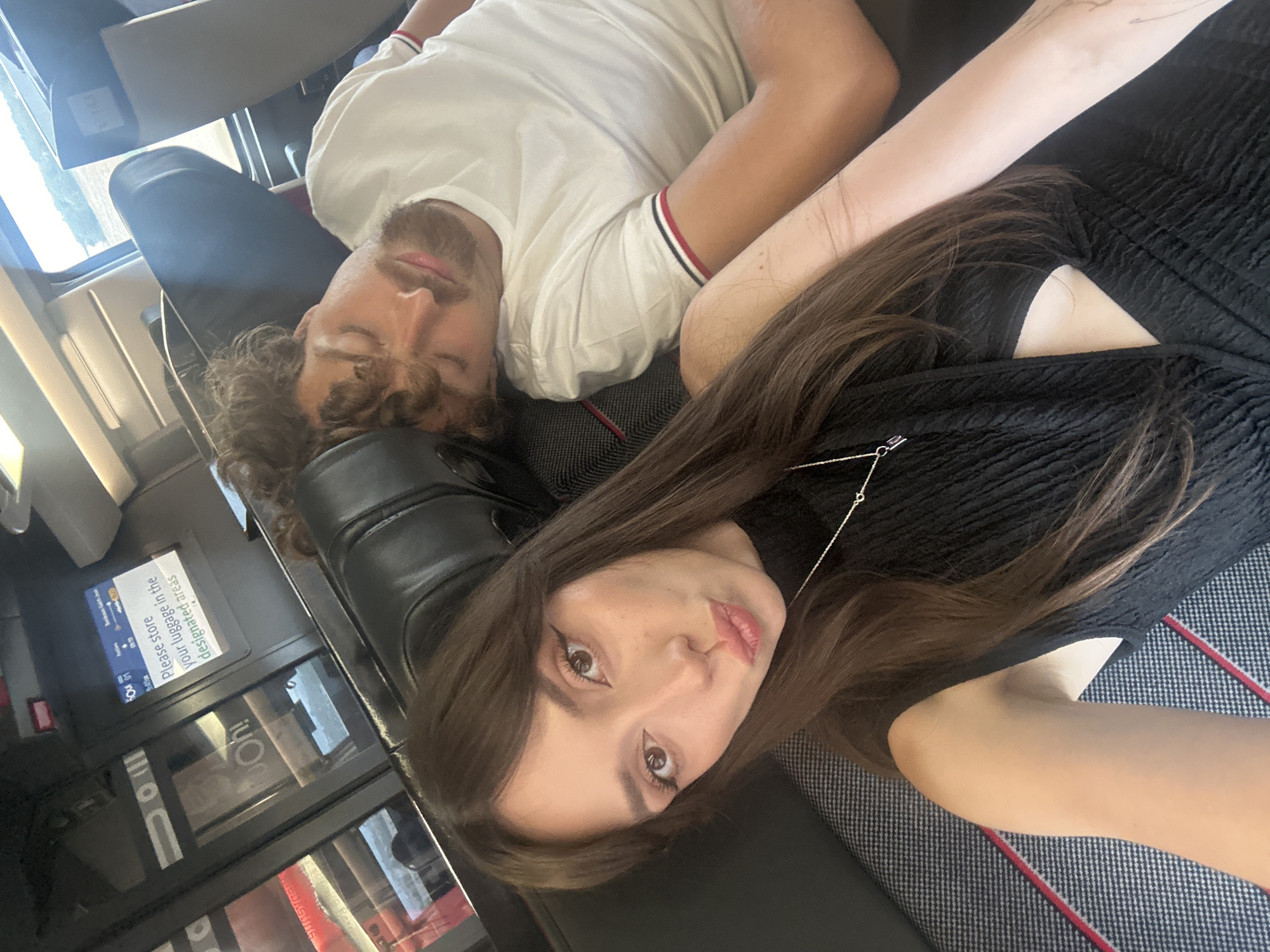 A selfie taken from a low angle shows a young woman with long brown hair and full lips puckered for a kiss. She is wearing a black, textured top with a cutout on the chest and a delicate silver necklace. Her left arm is extended to capture the shot. To her right, a man with curly brown hair and a beard sleeps with his head resting on a dark headrest. He is wearing a white t-shirt with red and blue stripes on the sleeve. They appear to be on a train, evidenced by the seats and a digital screen in the background displaying a message about storing luggage. The environment outside the window suggests a rural setting. The scene takes place on a train traveling through or near Fouqueure, France.