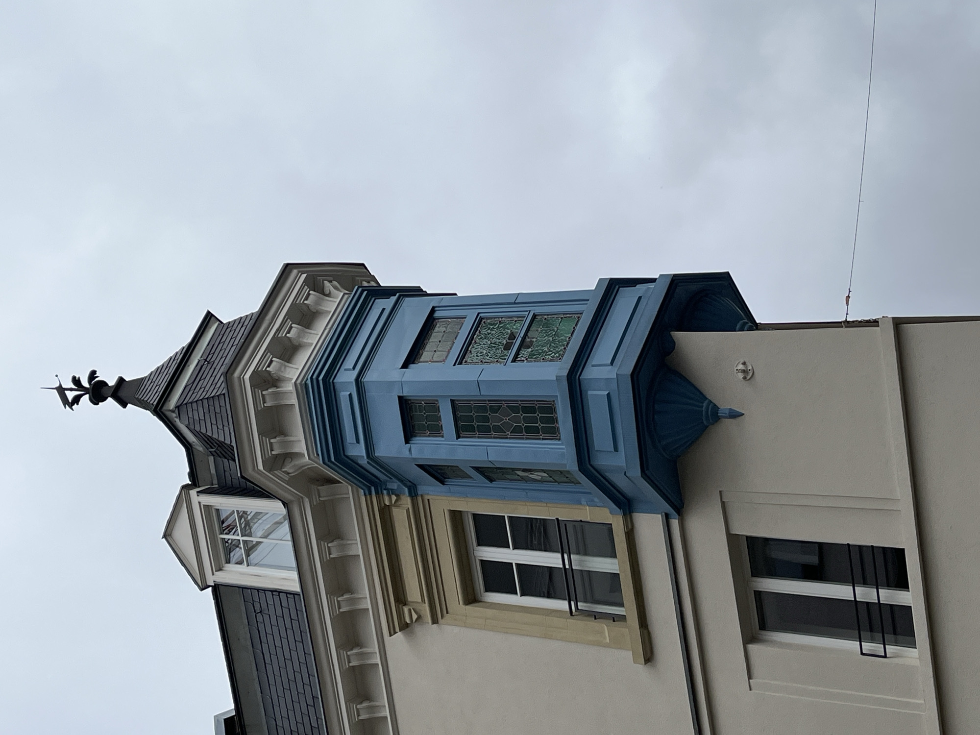 A low-angle shot depicts a section of a building in Luxembourg City, Luxembourg. The architecture is characterized by a mix of styles. The building comprises sections with diverse facade colors, including white and beige. A prominent blue or teal-colored bay window structure with decorative elements is visible. A dark, slate-tiled section with a small turret and ornate weather vane is attached. Standard rectangular windows are present on each level. The sky is overcast. A thin lightning rod is also visible.