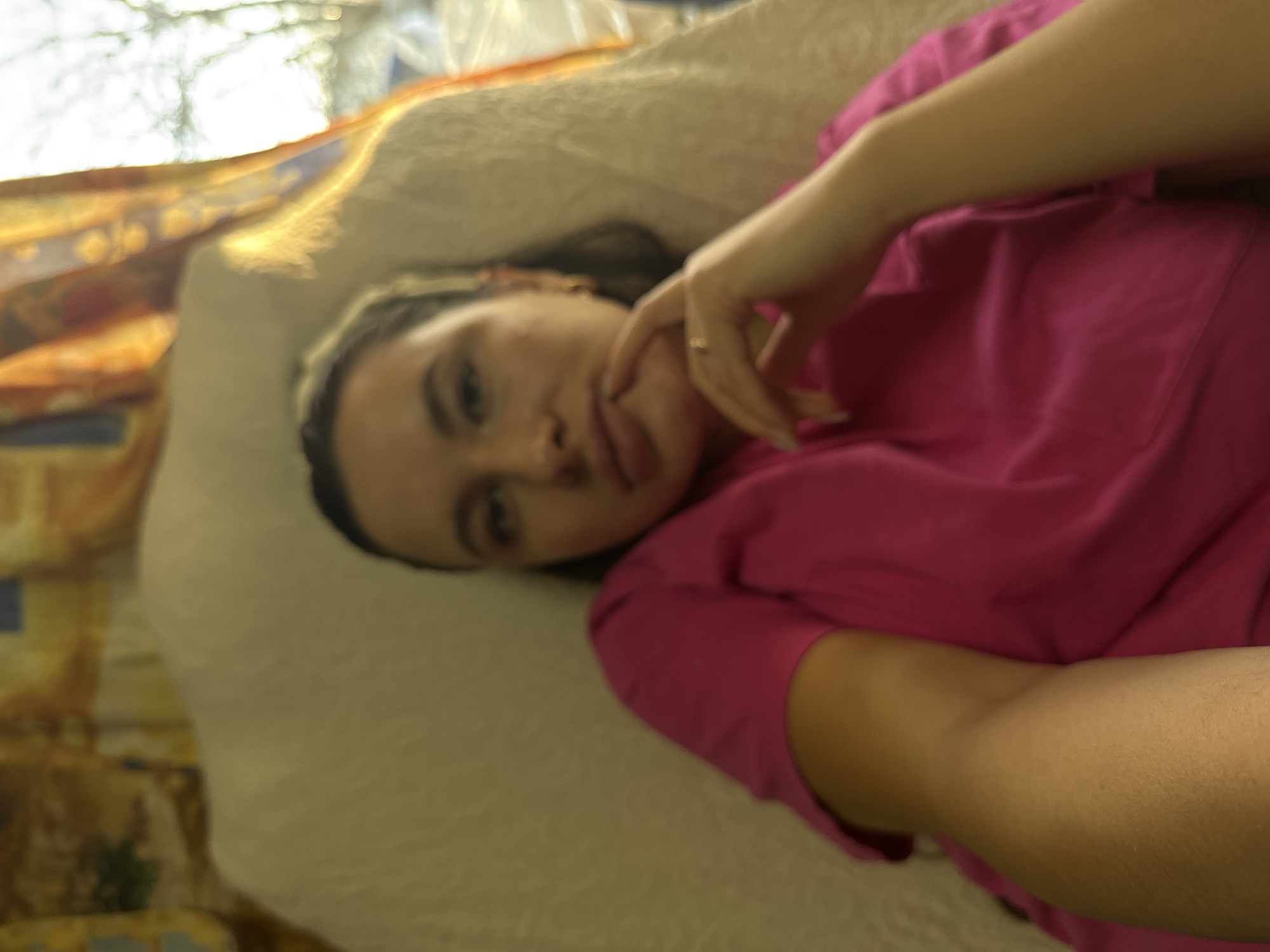 A young woman lies on a beige cushion or pillow, possibly in a domestic setting in or near Красный Маяк, Russia. She is wearing a bright pink shirt and has dark hair pulled back from her face. Her left hand is near her face, with a ring visible on one finger. The background includes a window and what appears to be a patterned fabric. The lighting suggests daylight.