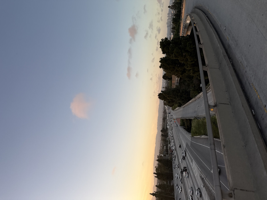 This image depicts an elevated view of a multi-lane urban freeway system at dusk, situated in Culver City, United States of America.
The foreground features a grey concrete overpass or ramp barrier, equipped with a metal guardrail containing yellow circular reflectors. The asphalt surface of this structure is visible, showing minor wear.
Below and to the left, a wide freeway with approximately six to eight lanes is visible, divided for opposing traffic flows. This freeway is heavily congested with numerous vehicles, primarily passenger cars and some trucks, moving in both directions. Headlights are visible on vehicles traveling away from the viewer, while the density of vehicles indicates slow movement.
To the right of the main freeway, a narrower, elevated road or ramp appears less utilized, flanked by dense, dark green conifer-like trees. Further right, another section of elevated road curves away from the viewer.
The background consists of rolling hills and a scattered urban landscape with various buildings. The sky transitions from a warm orange-yellow near the horizon on the far left, indicative of a sunset, to a pale blue overhead. A single prominent, diffuse pink cloud is centrally located in the upper sky, with a few smaller, fainter clouds scattered elsewhere. No individual people are discernible within the frame.