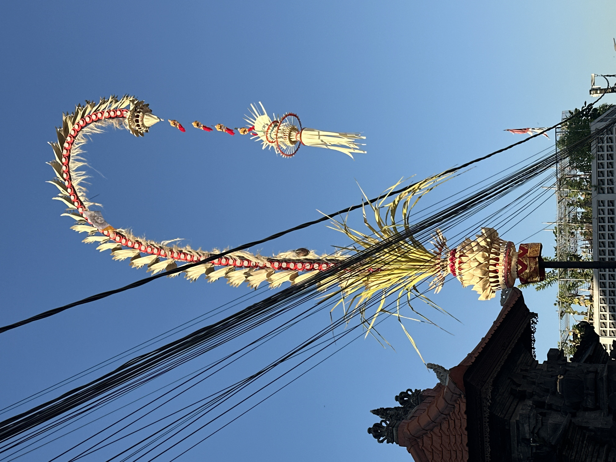The image depicts an outdoor scene in North Kuta, Indonesia, dominated by a bright blue sky. In the foreground, numerous black electrical wires crisscross the frame from left to right. On the left side of the image, a portion of a traditional Indonesian building with a red tiled roof and dark stone architecture is visible.
Rising from behind the building and extending upwards into the sky are elaborate, festive decorations. These appear to be of a spiritual or ceremonial nature, possibly related to a Balinese festival given the location. The main decoration is a tall, arched structure composed of overlapping, pale yellow or gold leaf-like elements outlined in red, resembling a dragon or serpent. White feathery strands are interspersed along this structure. Hanging from the curved upper portion of this decoration is a smaller, tiered ornament with white streamers. At the base of the main arched decoration, a crown-like structure made of similar pale yellow leaves and red accents is mounted on a dark pole, with a cluster of long, green fronds emerging from its top.
In the distance, a small red and white flag is visible on a pole, and beyond that, a modern building with a white patterned wall and greenery on its rooftop can be seen. The overall impression is one of a vibrant cultural display set against a clear, sunny sky, juxtaposed with the everyday infrastructure of electrical wires. No people are clearly visible in the image.