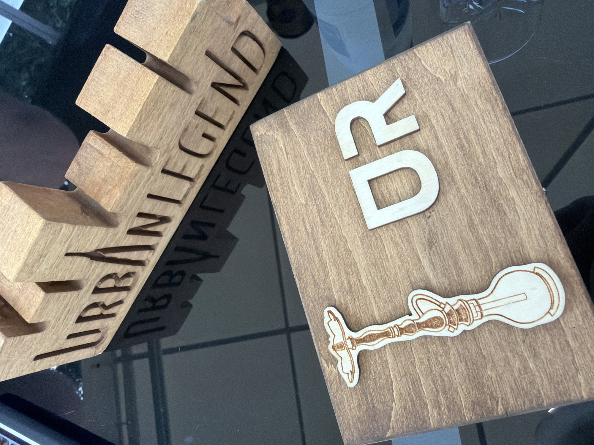 The scene is located in Sofia, Bulgaria. The image depicts two wooden decorative items resting on a dark, highly reflective surface, likely a tabletop.
In the upper portion of the frame, a horizontal wooden block is positioned, featuring the words "URBAN LEGEND" carved deeply into its front face. The block's upper edge is cut into an irregular, stepped profile, resembling miniature city buildings. The wooden material exhibits a light brown, possibly maple or similar light-toned wood, with visible grain. Its reflection is clearly visible on the dark surface below.
In the foreground, slightly to the lower left, a flatter, rectangular wooden plaque is visible. This plaque is made of a darker, stained wood, displaying a distinct wood grain pattern. Attached to its surface are two distinct wooden elements:
1. On the left, a laser-cut or carved wooden silhouette of a stylized hookah (shisha pipe) is affixed. The outline is clearly defined by darker burnt edges, and internal details like the base, stem, and bowl are etched onto its surface.
2. To the right of the hookah silhouette, two individual wooden letters, "U" and "R," are mounted. These letters are of a lighter wood tone, contrasting with the darker plaque, and appear to have a slightly raised, dimensional quality.
The reflective dark surface shows clear, inverted reflections of all the wooden objects. In the far background, out of focus, diffused light sources and indistinct green elements, possibly foliage, are discernible, suggesting a window view or an illuminated area beyond the immediate setup. A partial view of a dark, oblong object, possibly an electronic device, is visible at the very top left edge of the frame. No individuals are present in the image.