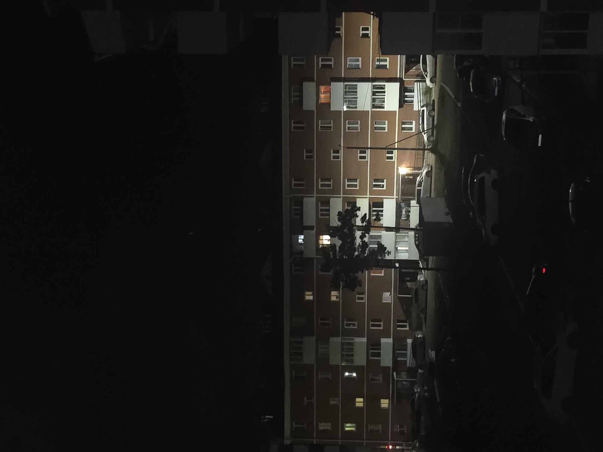 The image captures a nighttime view of a multi-story apartment building in Ulan-Ude, Russia. The building is illuminated by scattered lights from its windows, suggesting occupancy. A tall tree is visible in front of the building, partially obscuring some of the windows. In the foreground and middle ground, several cars are parked in a courtyard or parking lot area, some of which are partially illuminated by streetlights or the building's lights. The overall scene is characteristic of a residential area at night, with a sense of quietude. The architecture of the building appears to be typical of Soviet-era housing blocks. The sky is dark, indicating it is well past sunset.