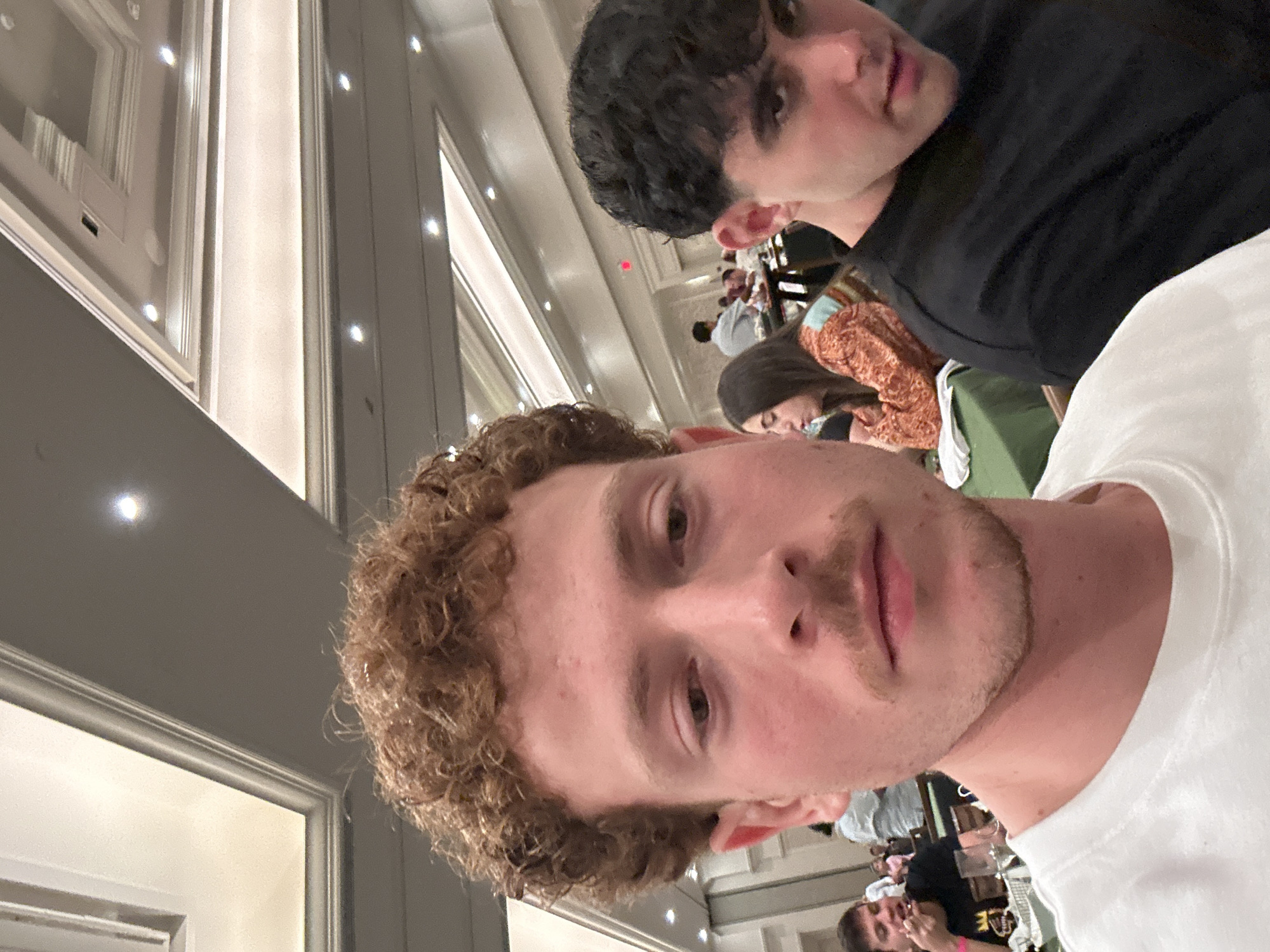 Two males are in the foreground of a vertically oriented, angled, indoor photograph. The male in the immediate foreground has light skin, a light mustache, and curly, reddish-brown hair. He is wearing a white shirt. Behind him, another male with dark hair is wearing a black shirt. Several other people are out of focus in the background, sitting at tables in what appears to be a dining hall. The location is in the city of Aventura, United States of America.