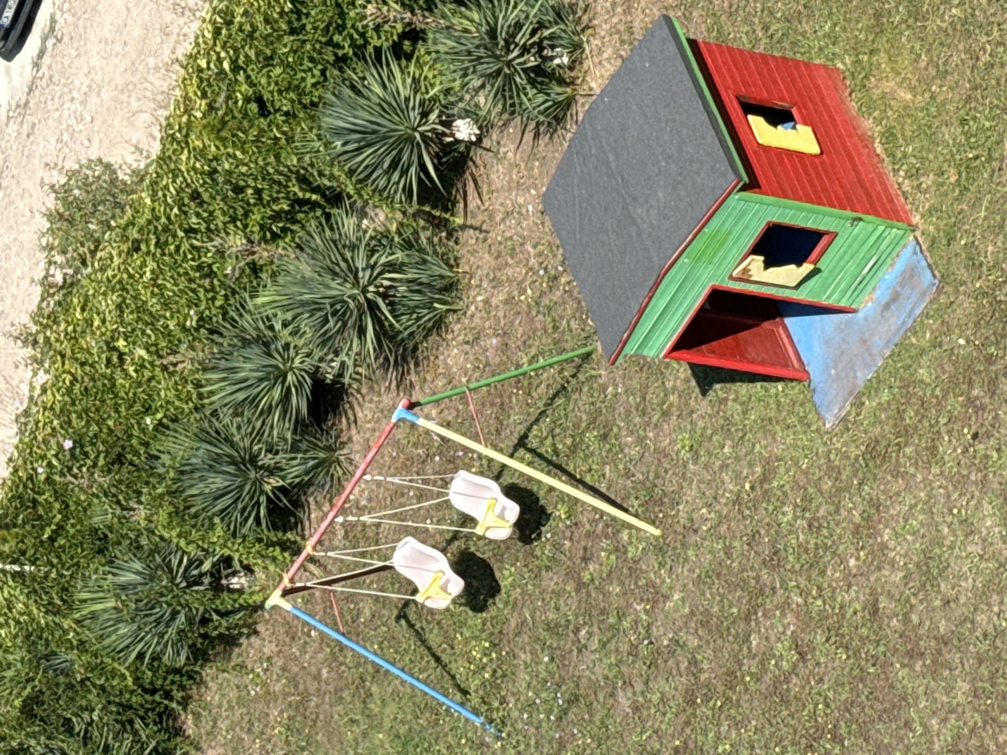 An aerial, high-angle, full shot captures a small, weathered, red and green children's playhouse and a swing set situated in a grassy, uneven, dirt area with scattered weeds and small yellow flowers. The playhouse, with a grey, flat roof, features a red, open doorway and two small windows, one on the front and one on the side, both with yellow inserts, one of which appears broken. The swing set, with blue, red, and yellow uprights and crossbars, supports two light-colored swings, each with yellow restraints. Dense green foliage, likely ivy, covers a textured wall or embankment behind the play area, interspersed with spiky green plants. The scene is located in Pomorie, Bulgaria, on a sunny day.